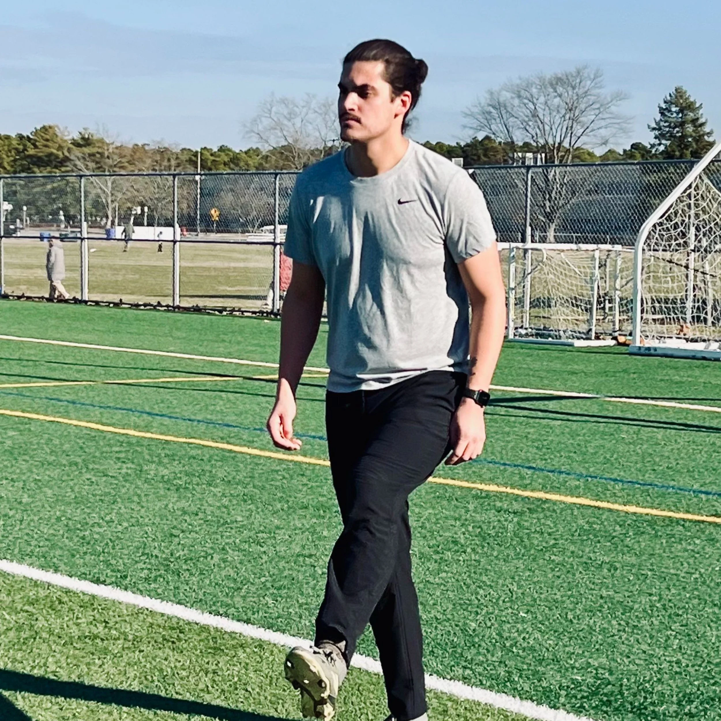A young man in a gray T-shirt and black pants walking on a soccer field with artificial grass. He has dark hair tied back and is wearing a watch on his left wrist. The background features a fence, trees, and other people in the distance.