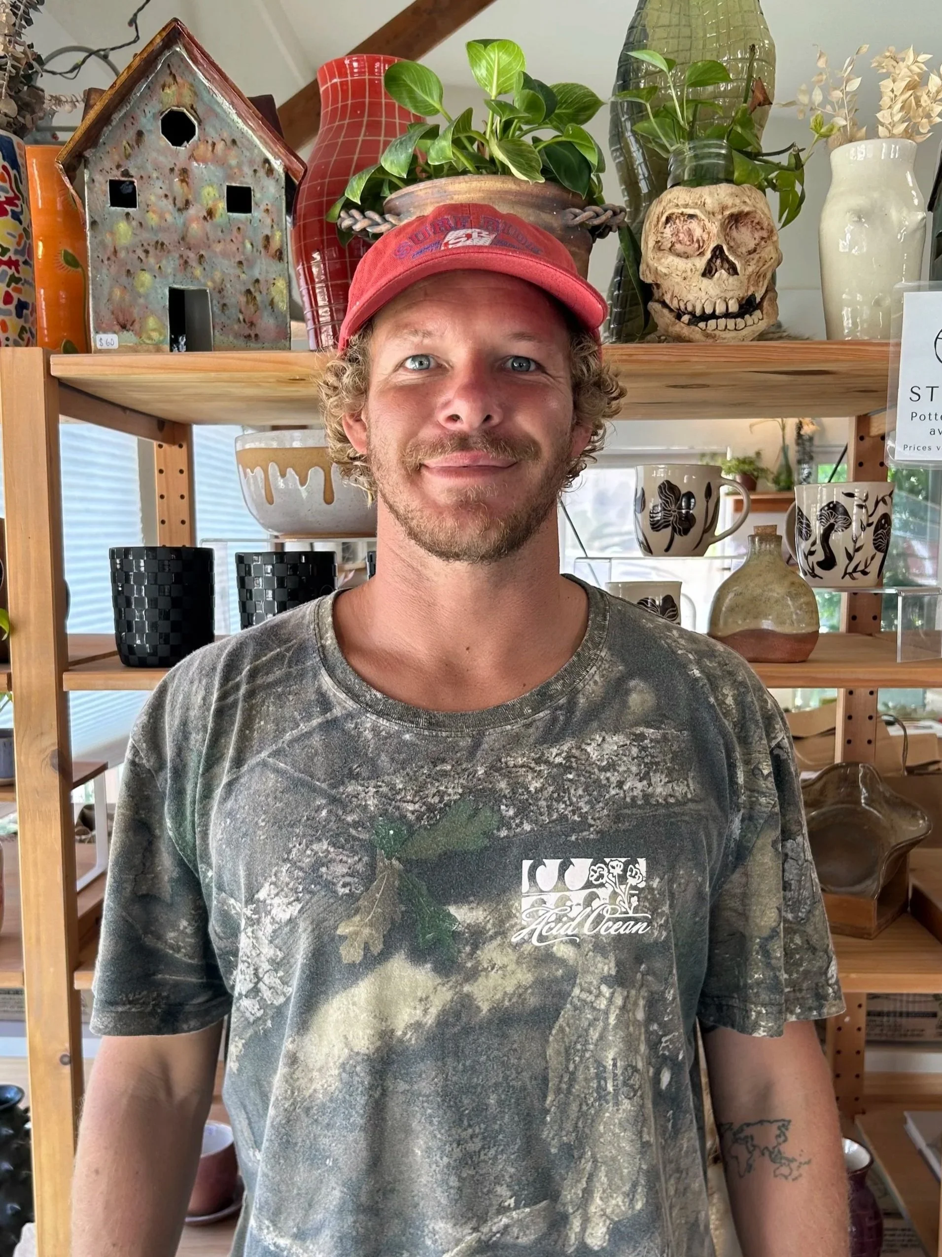 A man with curly blond hair and a beard, wearing a red cap and a tie-dye t-shirt, standing in front of shelves with decorative items including pottery, a skull, and plants.
