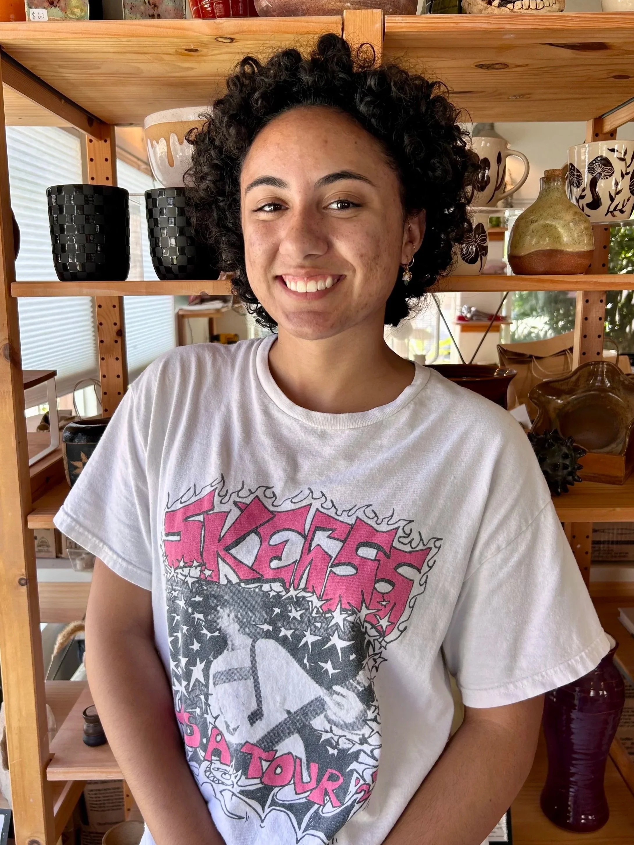 A smiling young woman with dark, curly hair stands in front of a wooden shelf filled with various ceramic and pottery items. She is wearing a white T-shirt with a graphic design.