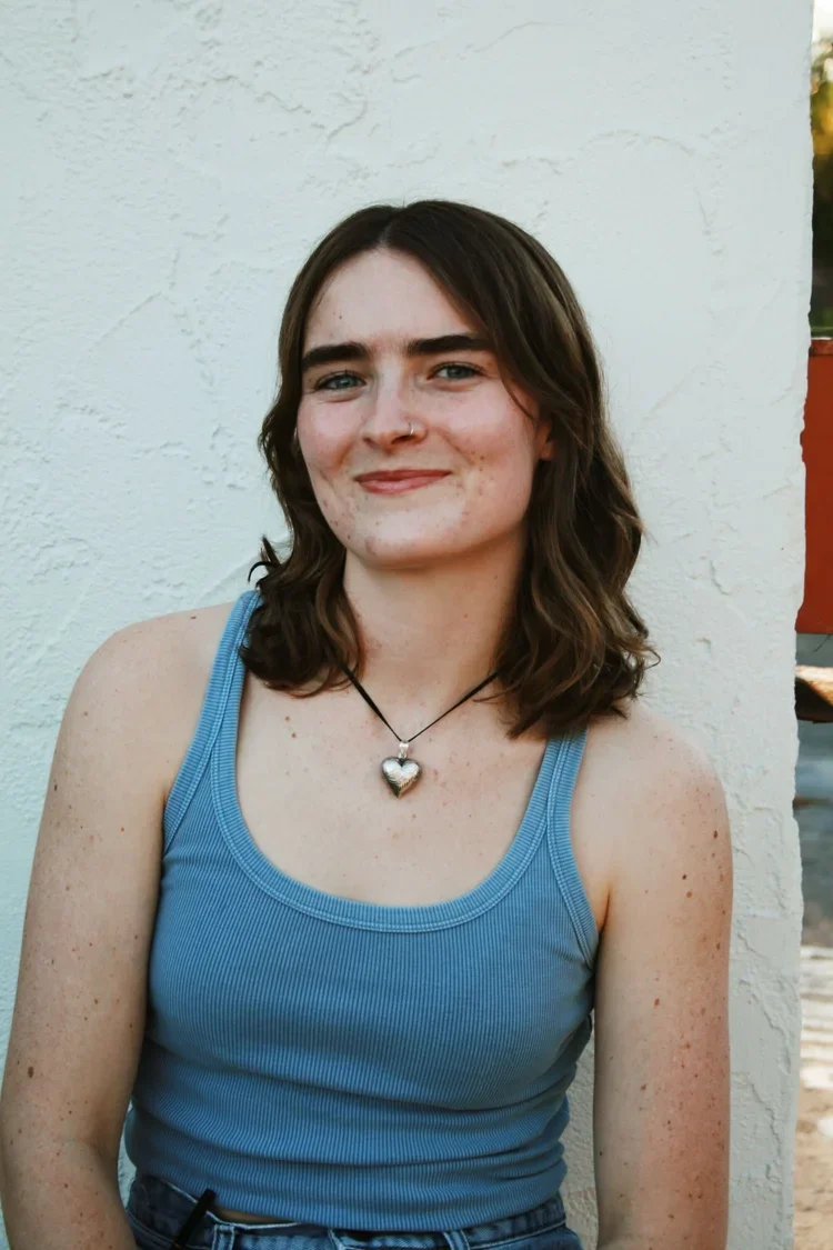 A young woman with shoulder-length brown hair and freckles, smiling, wearing a light blue tank top, a black necklace with a heart-shaped pendant, and a nose piercing, sitting against a white textured wall outdoors.