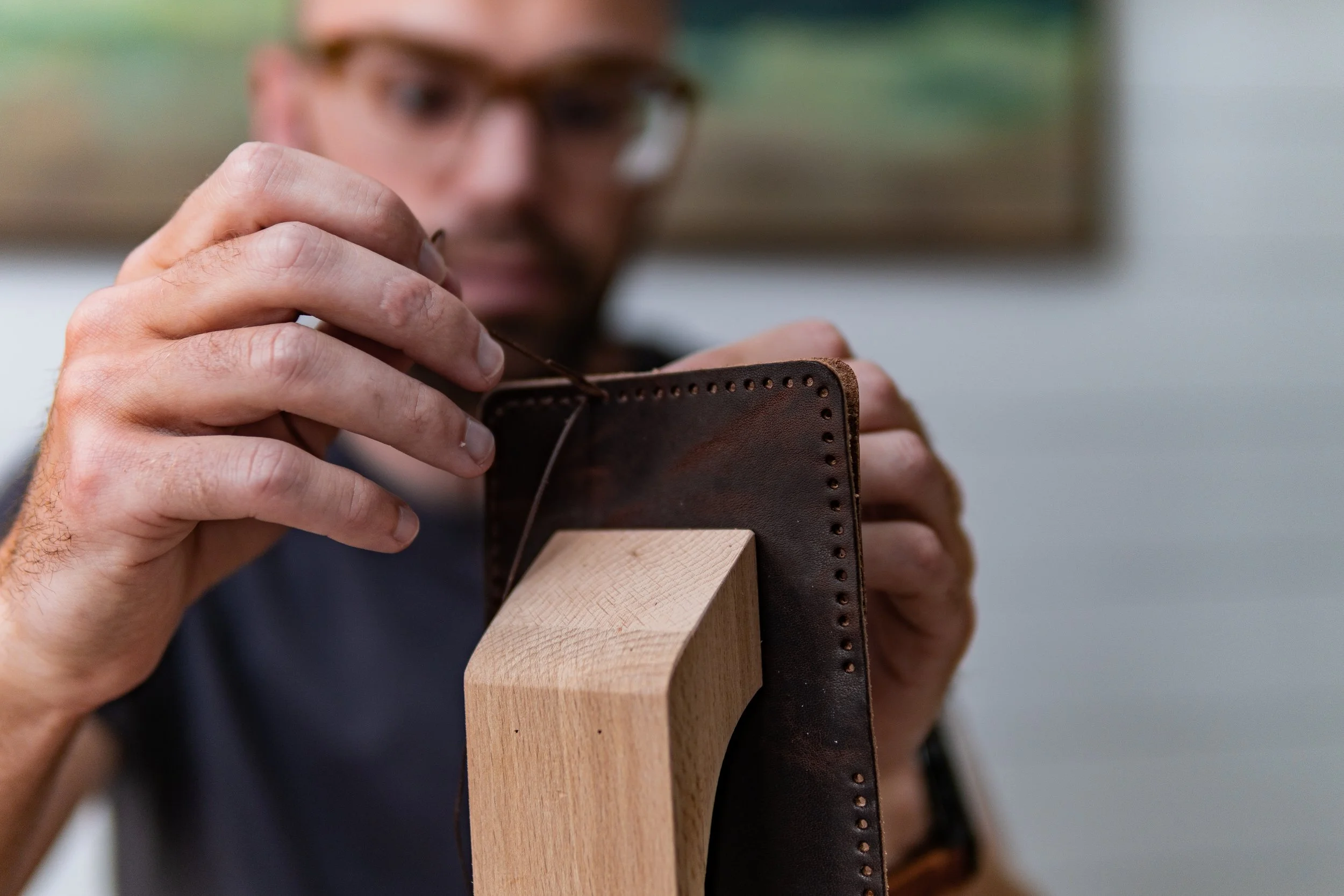 A craftsman wearing glasses is stitching a leather piece with a wooden block to support it.