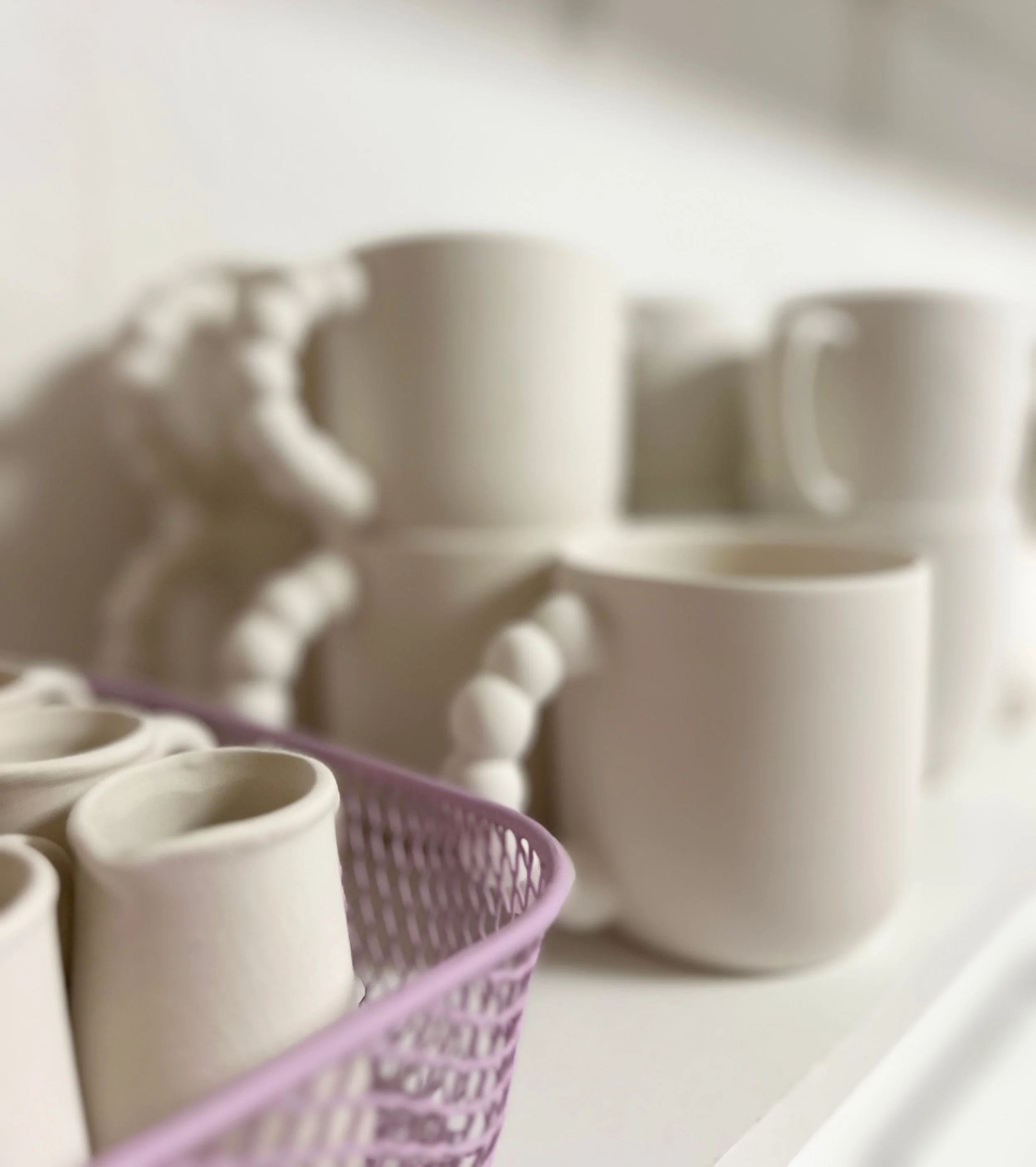 Weiße Kaffeetassen, einige in einem lilafarbenen Plastikkorb und andere auf einem weißen Regal, alles unscharf im Hintergrund.