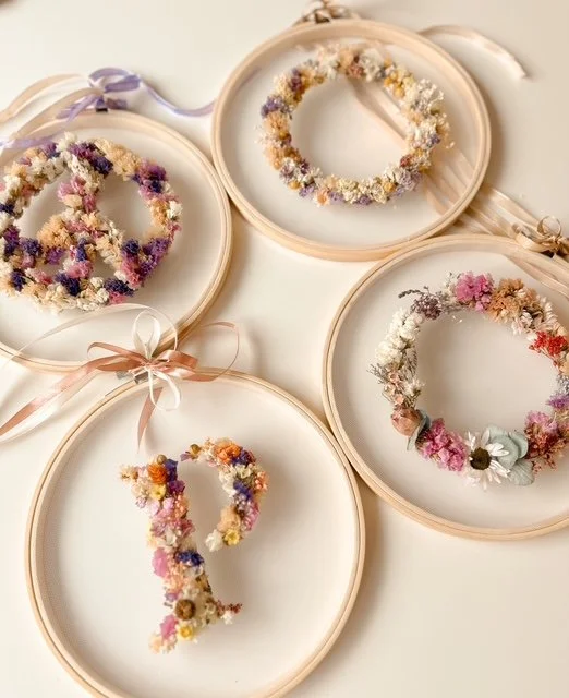 Vier kreisförmige Blumenkränze in Stickrahmen, dekoriert mit getrockneten Blumen in Pastellfarben, auf eine weiße Oberfläche gelegt.