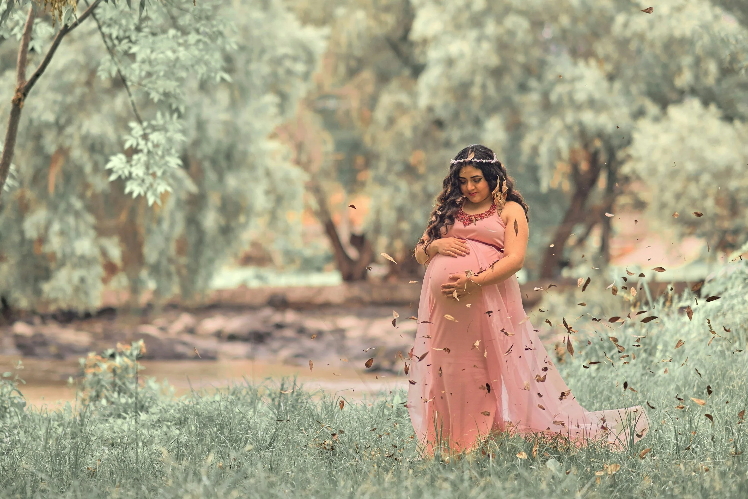 Pregnant woman in a pink dress holding her belly in a natural outdoor setting with trees and grass, surrounded by falling leaves.