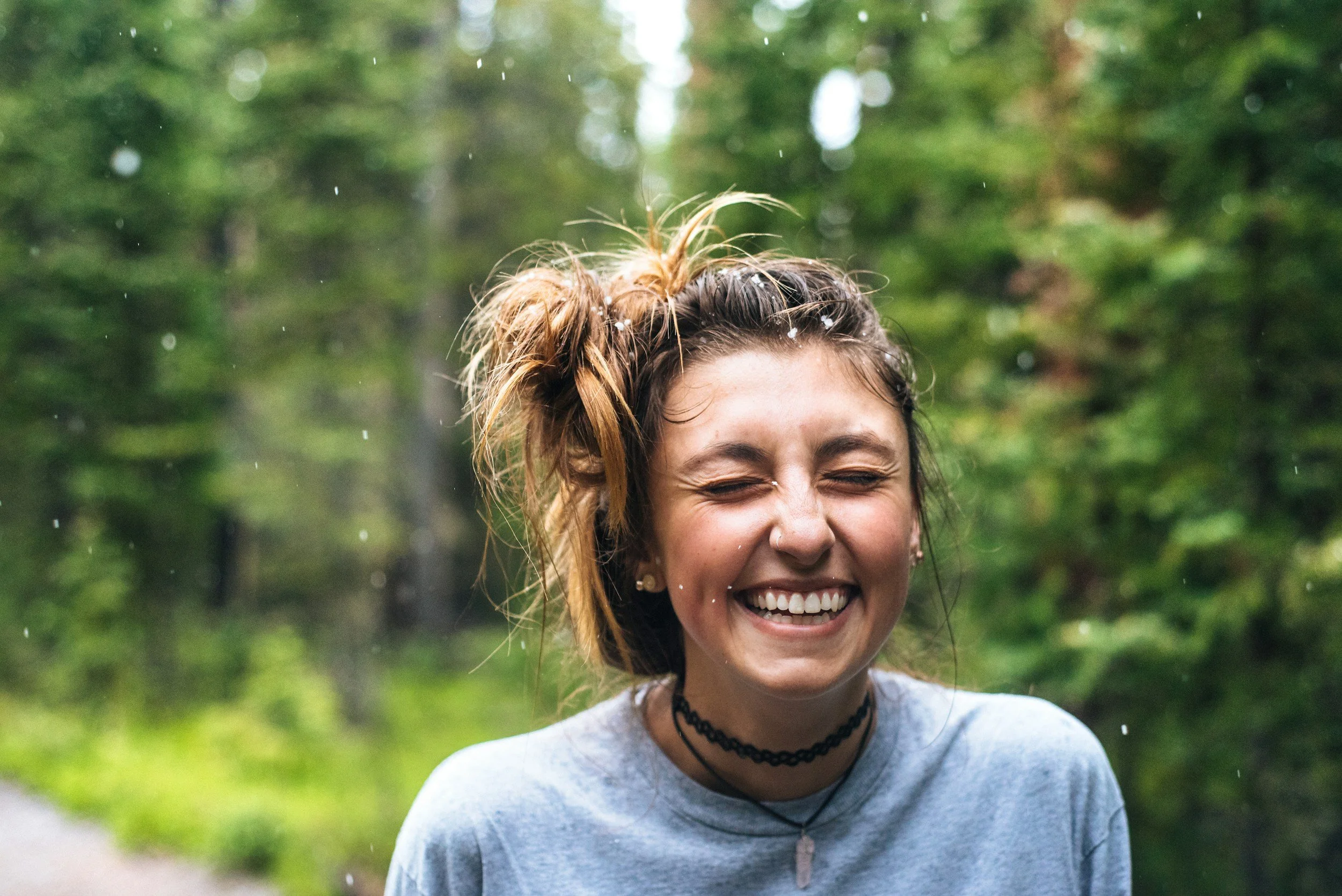A young woman with wet hair smiling and laughing in a wooded outdoor setting.