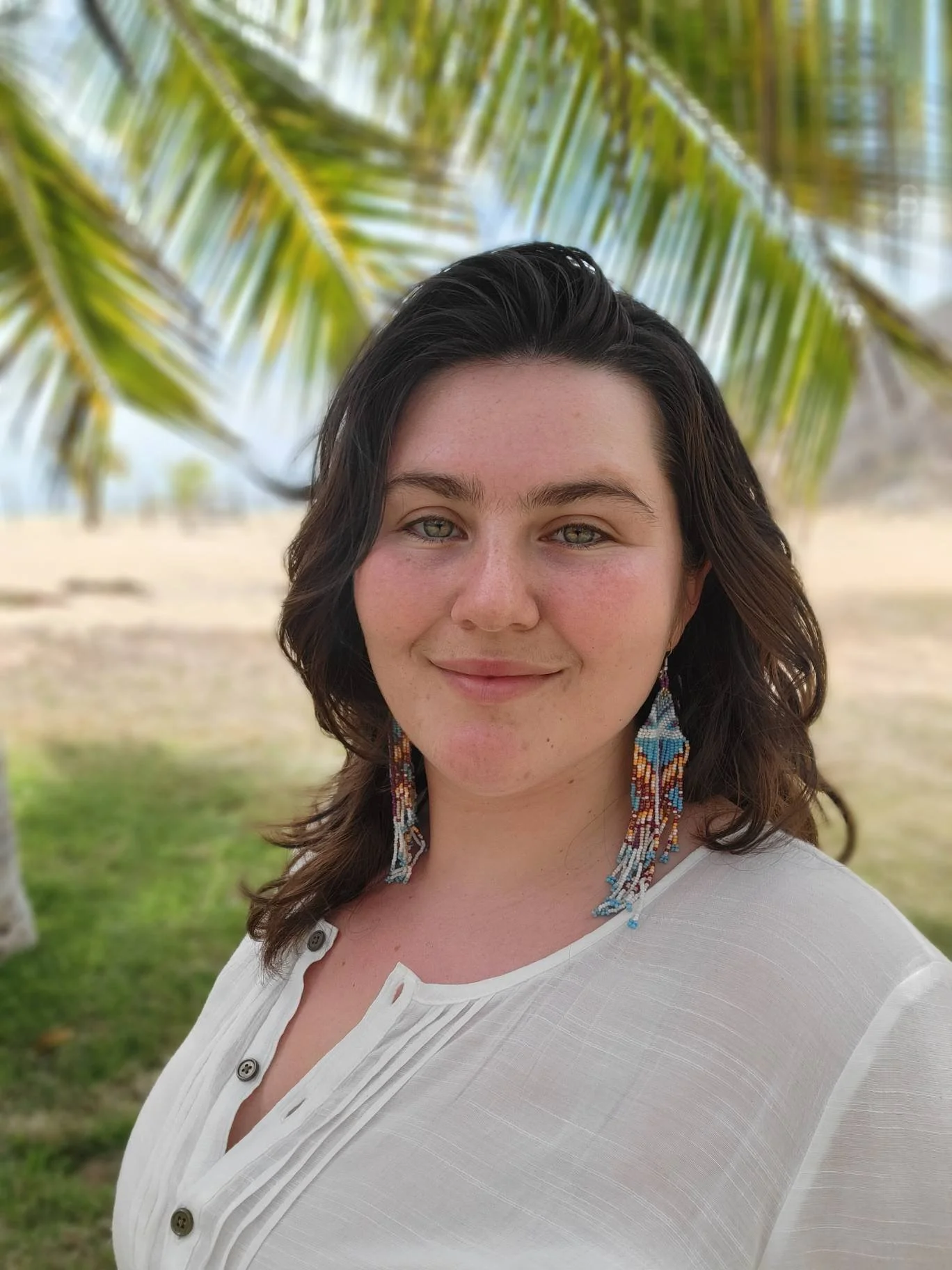A young woman smiling outdoors with palm tree leaves in the background, wearing beaded earrings and a white buttoned blouse.