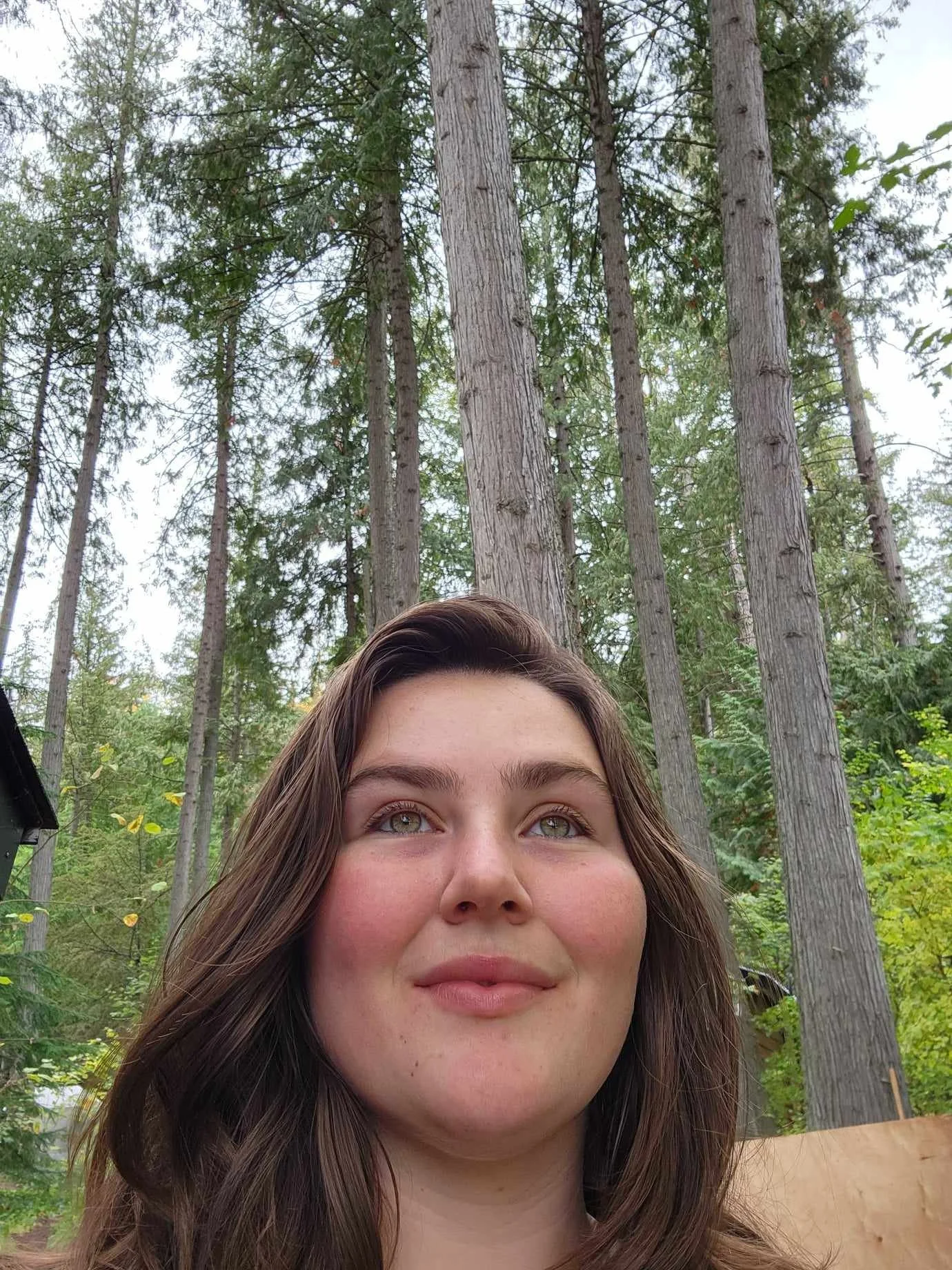 A young woman with shoulder-length brown hair and light makeup, outdoors in a wooded area with tall trees and green foliage, looking slightly upwards with a calm expression.