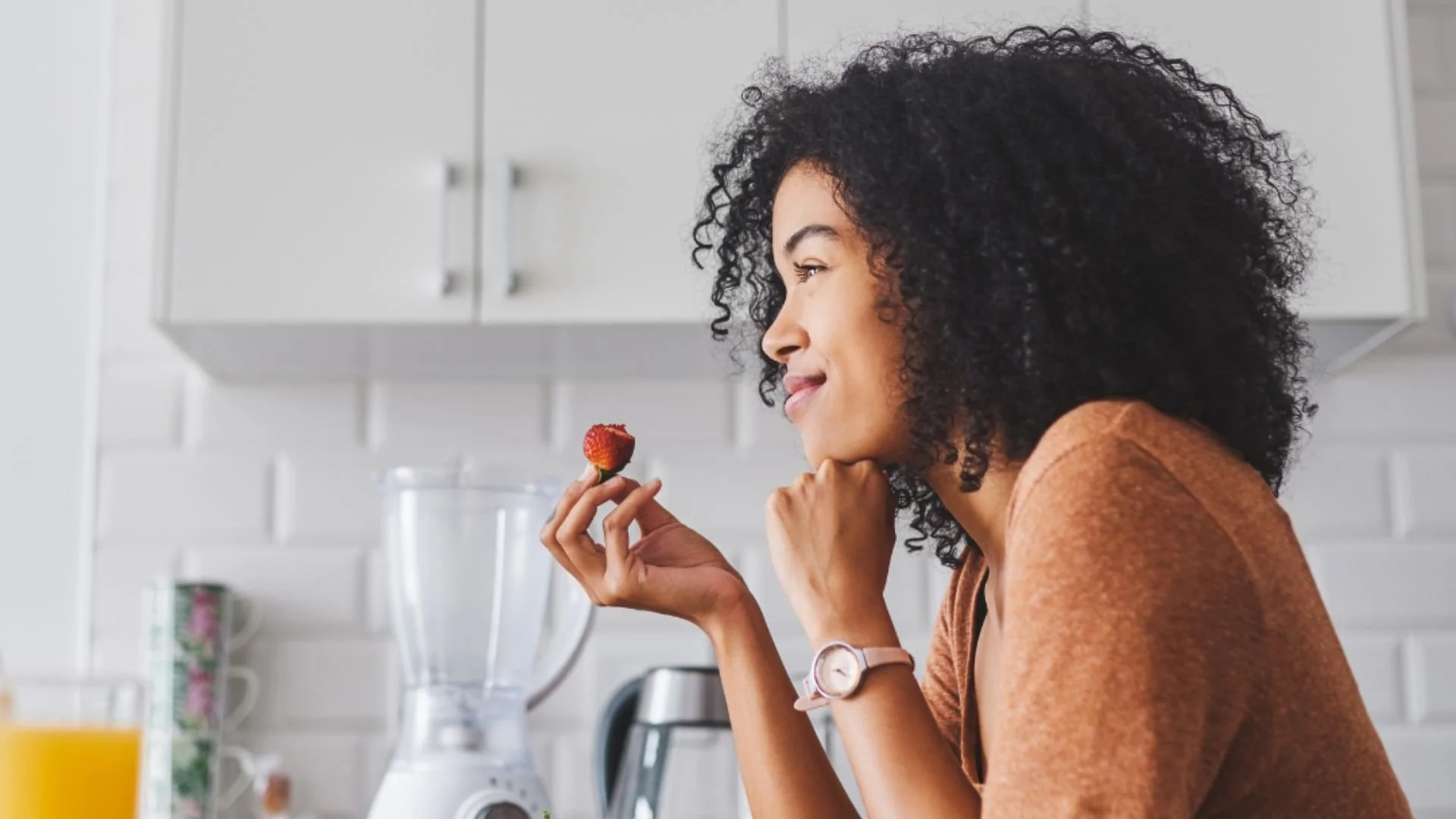 A woman with curly black hair holding a strawberry in her right hand, smiling and looking at it in a kitchen setting.