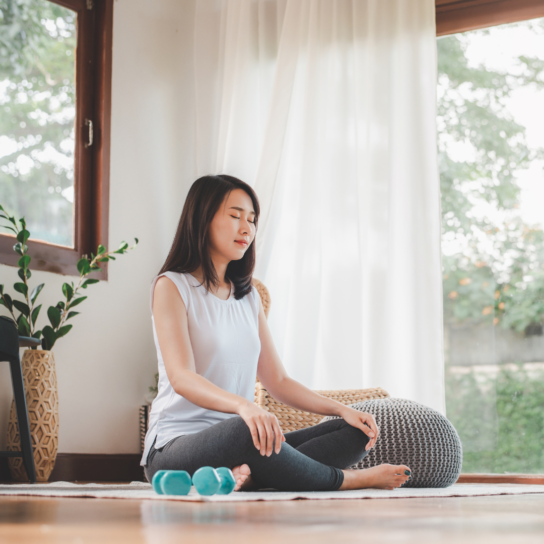 Woman practicing meditation on a rug in a bright room with sheer curtains, a plant, and a window showing trees outside.
