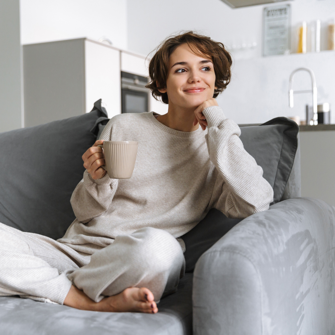 A young woman with short brown hair sitting on a grey couch, holding a beige mug, smiling thoughtfully, in a cozy modern kitchen.
