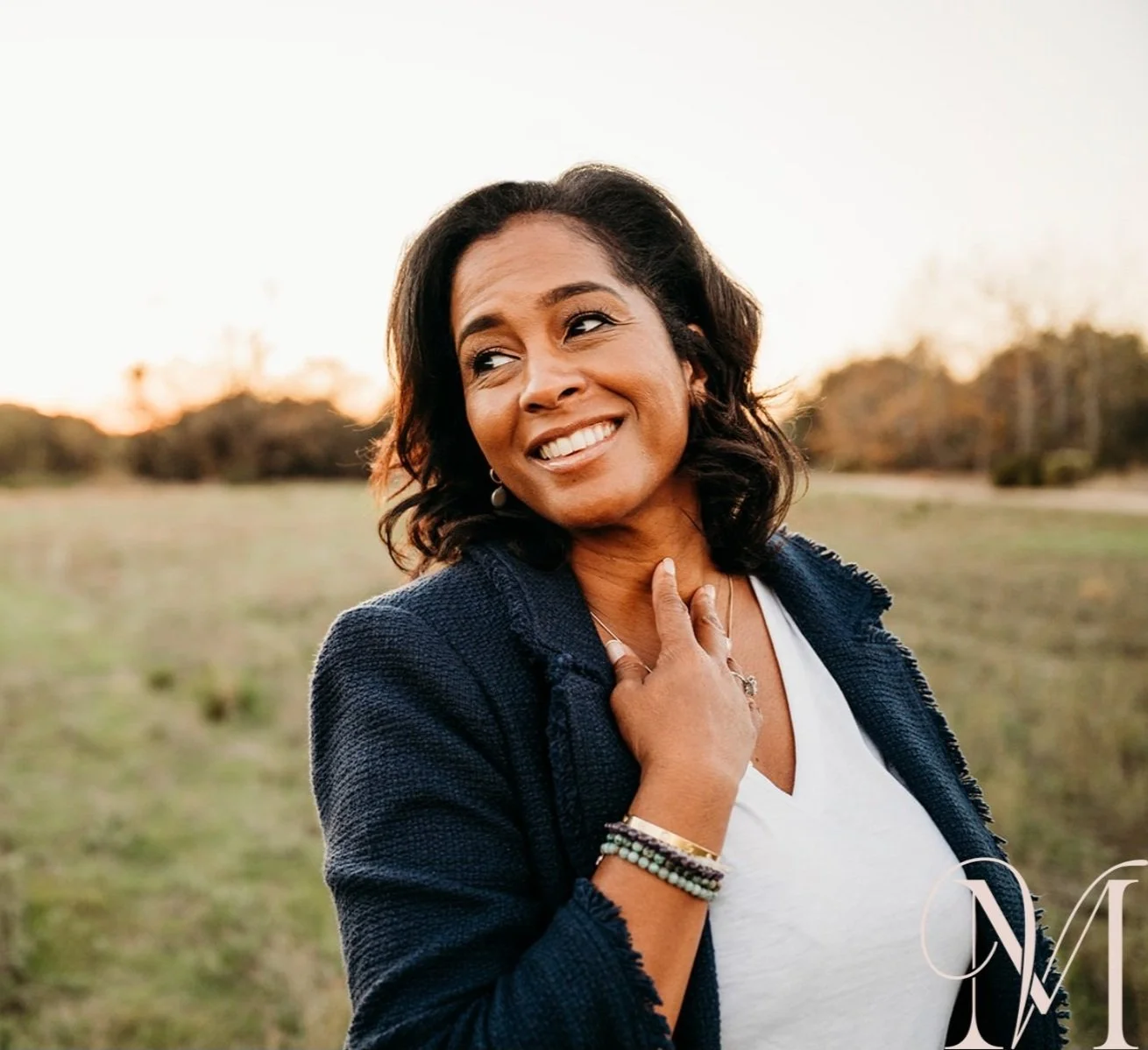 A smiling woman with shoulder-length dark hair, wearing a navy blazer and white top, standing outdoors in a field at sunset, with one hand on her chest and jewelry on her wrist and fingers.