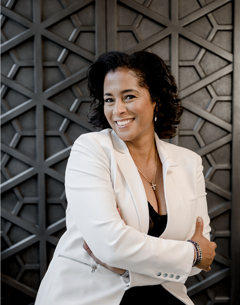 A woman with short curly black hair smiling, wearing a white blazer over a black top, standing in front of a geometric patterned black wall.