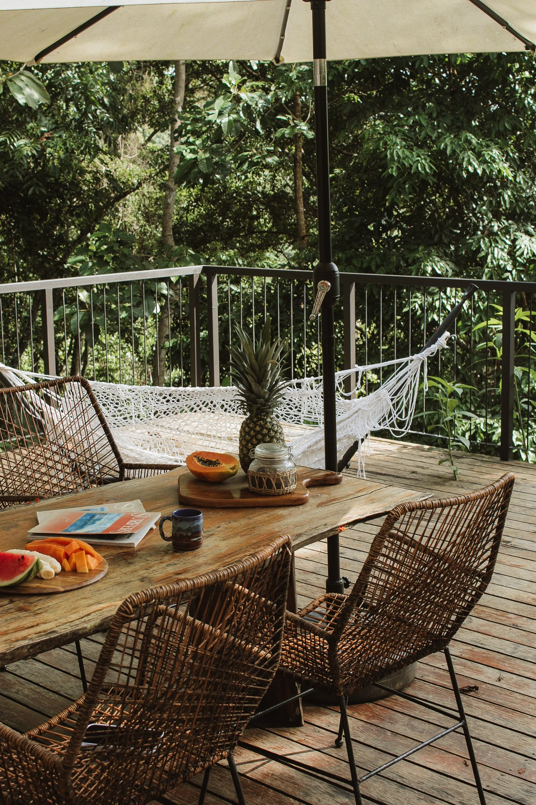 Outdoor wooden deck with a rustic table set for breakfast, featuring a pineapple, papaya, watermelon, and a magazine, surrounded by wicker chairs, a hammock, and a large patio umbrella, with lush green trees in the background.
