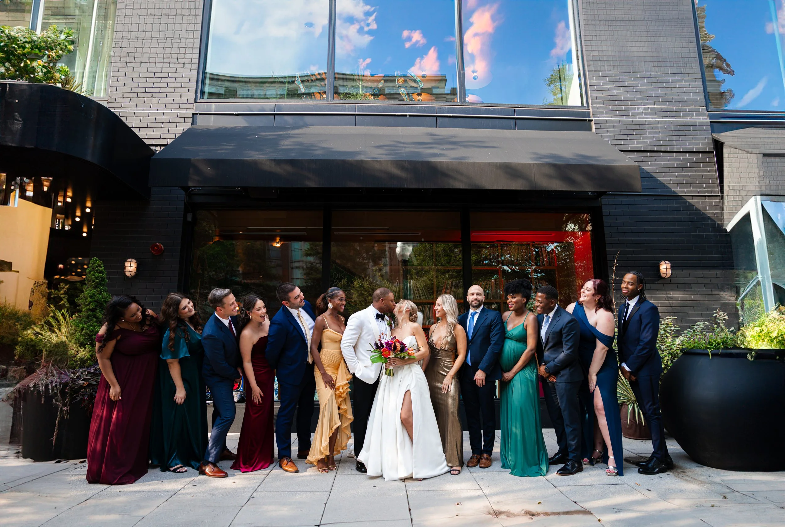 A diverse group of people in formal attire, including a bride and groom, gathered outside a modern building, celebrating a wedding.