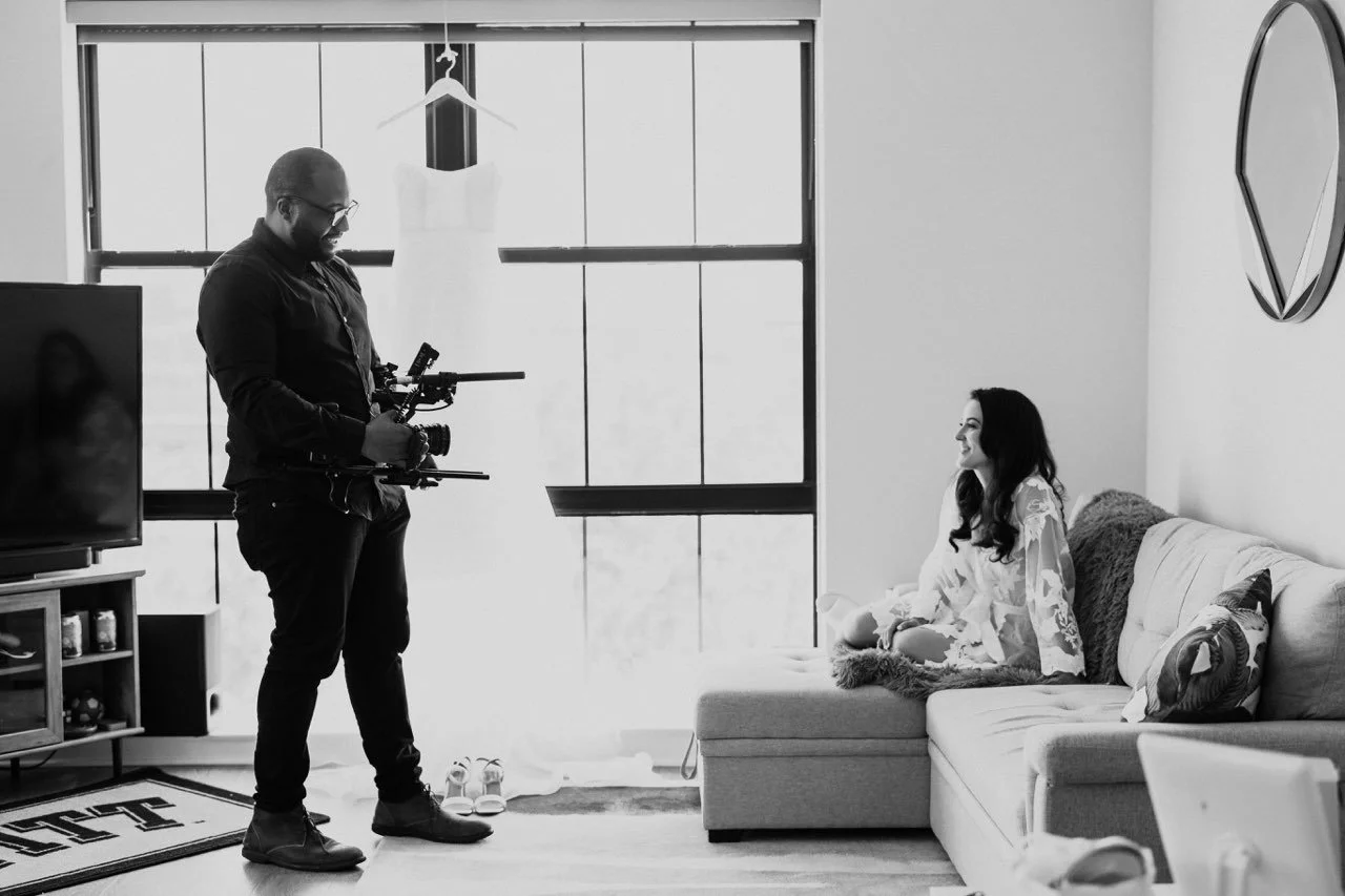 A man filming a woman sitting on a sofa in a living room during a photoshoot.