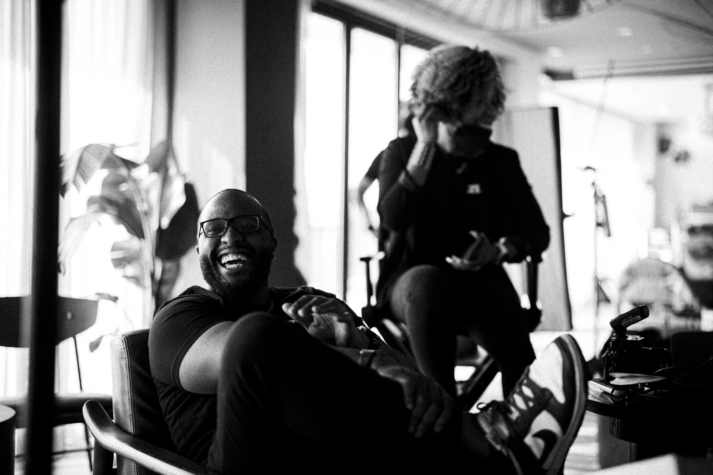 A black-and-white photo of a man with glasses, a beard, and a short haircut, smiling and sitting comfortably in an indoor space. Another person with curly hair and glasses, sitting on a chair and looking at their phone, is in the background. The setting appears to be an informal meeting or casual social gathering near large windows letting in natural light.