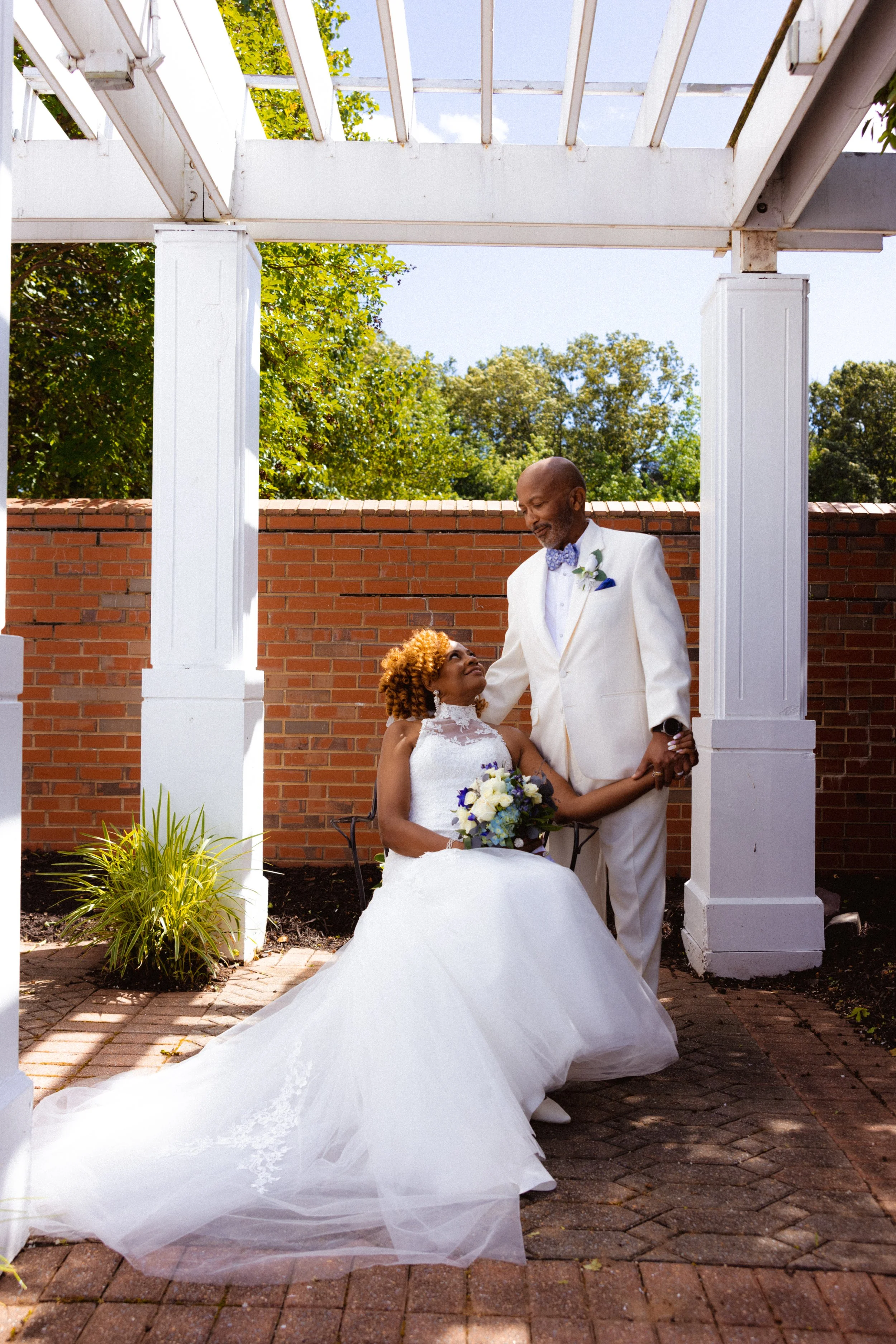 A bride and groom share a loving moment outdoors under a white wooden pergola. The bride is seated, wearing a white wedding gown and holding a bouquet of white and purple flowers. The groom, dressed in a white suit with a bow tie, stands beside her, 