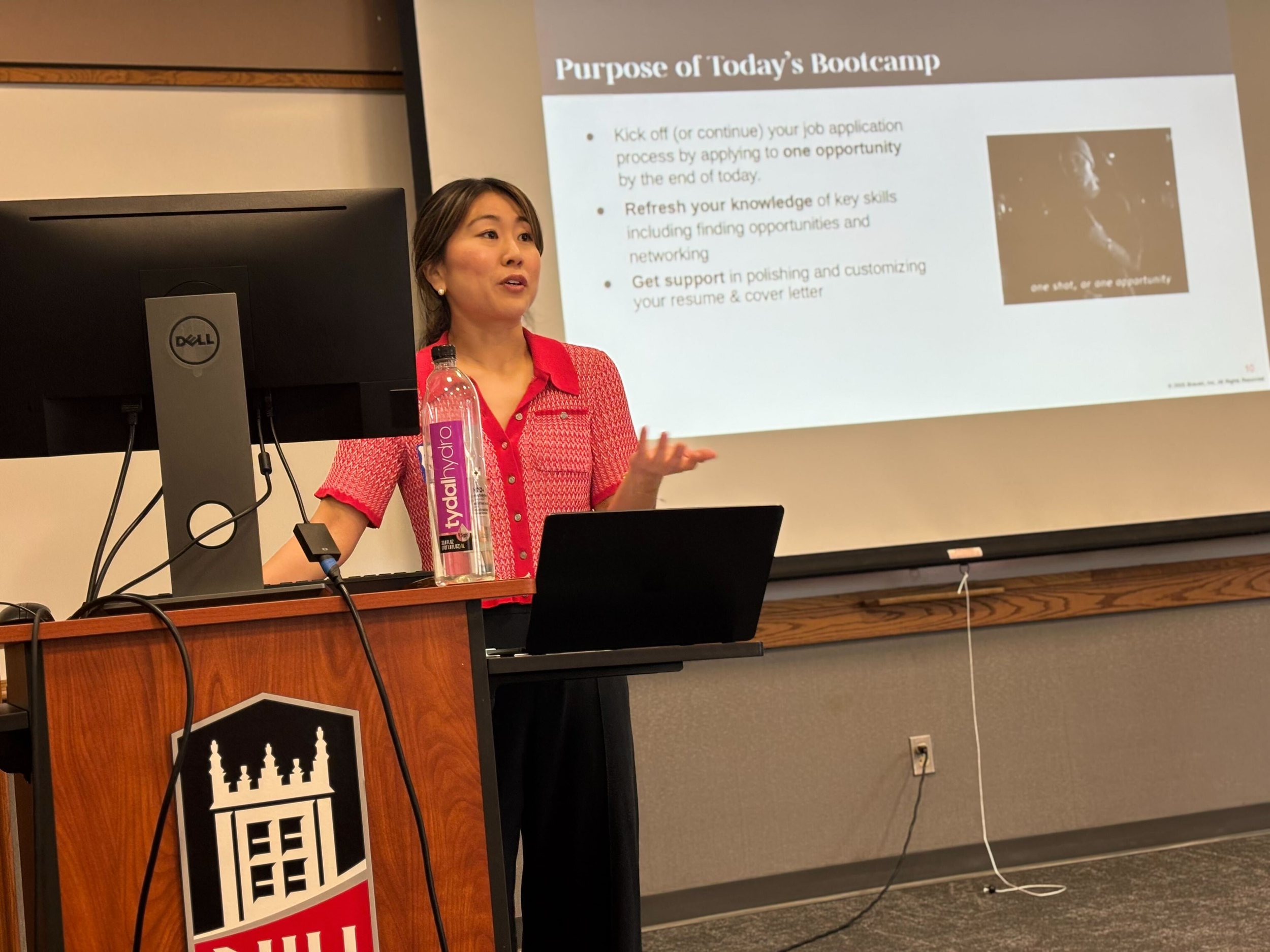 A woman in a red shirt giving a presentation in front of a slide that reads 'Purpose of Today's Bootcamp'. She is standing next to a lectern with a sign of a city skyline. There is a water bottle on the lectern. The background includes a computer monitor and a wall with a power outlet.