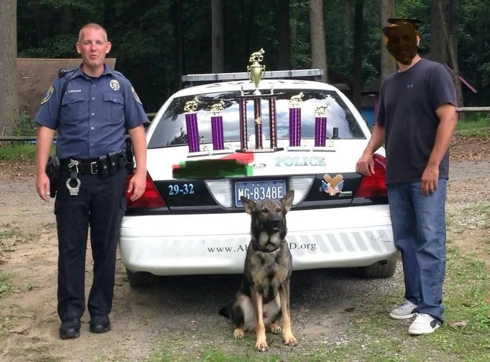 A police officer and a civilian stand on either side of a police car with a German Shepherd sitting in front, outdoors in a wooded area. The trunk of the car displays trophies and awards.
