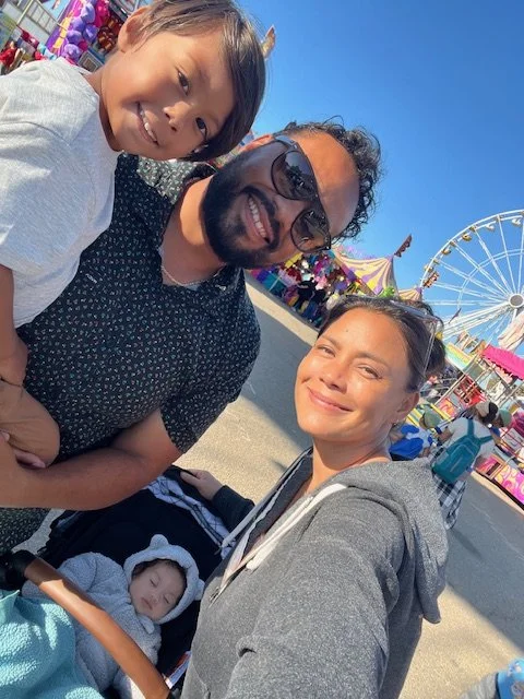 Group photo of two women, a man, a young girl, and a baby at a fair with a Ferris wheel in the background, sunny weather.