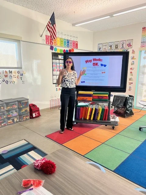 Women standing in a kindergarten classroom in front of a tv that reads Play Monde: On. She is wearing purple sunglasses, black pants and a white shirt with blue organic spots. Women's hands are up and she is talking.