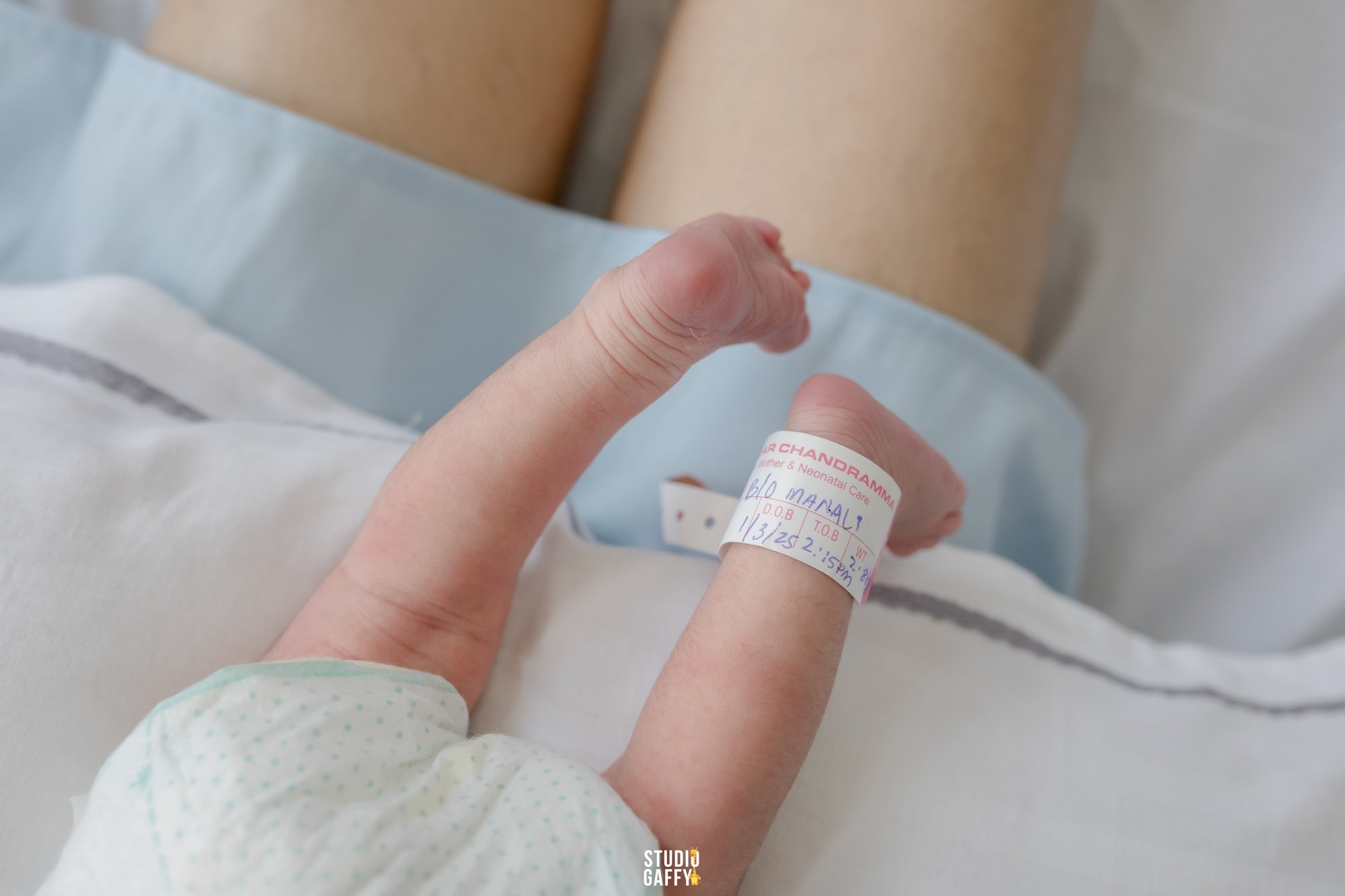 A close-up shot of a person's hand with an IV bandage, holding their first finger. The person is lying in a hospital bed, visible through the hospital gown's sleeve. The bandage has a medical label with handwritten details, including the name 'BLO MA