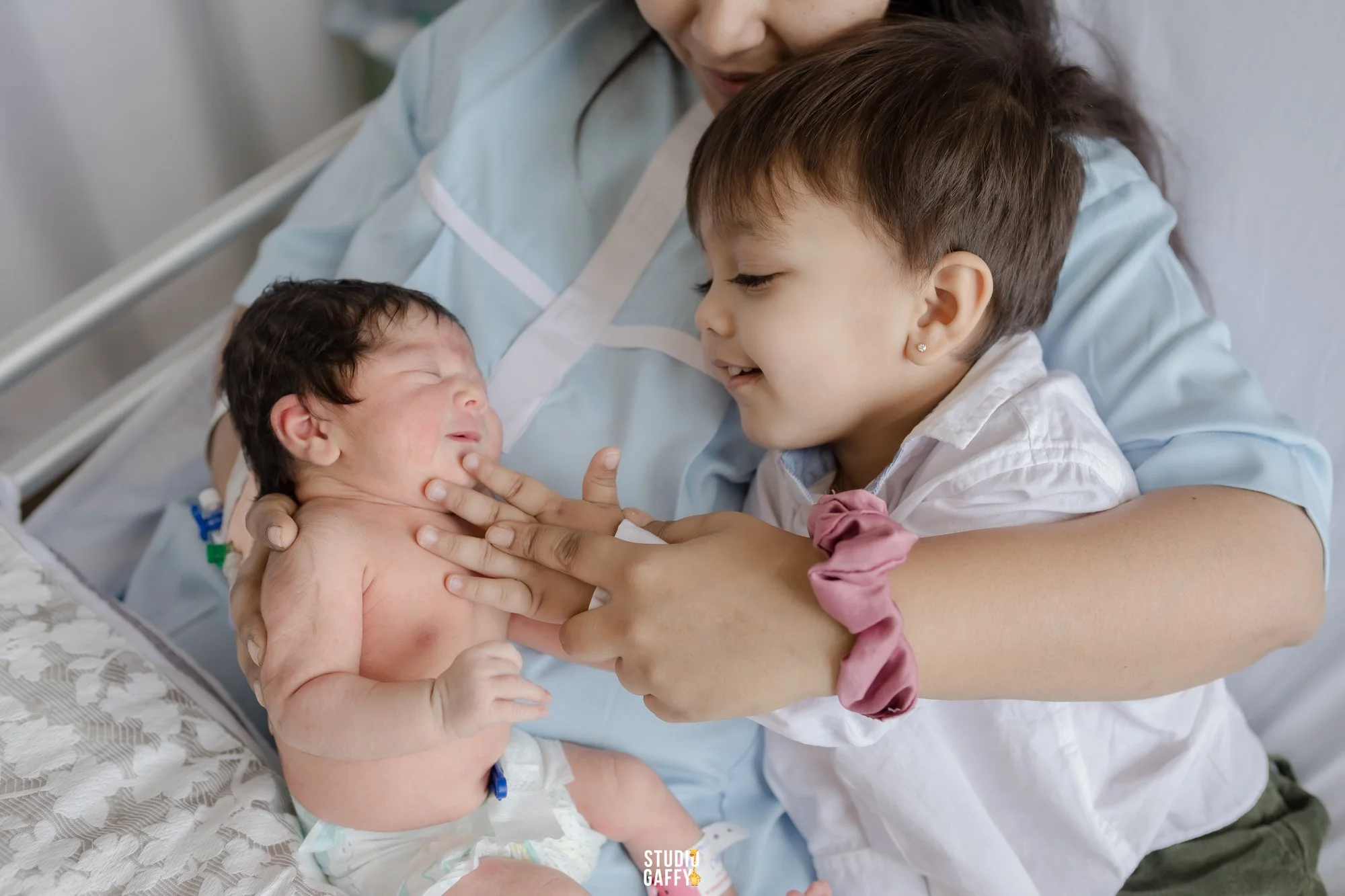 A woman in hospital gown holding a newborn baby with a woman and a young girl looking at the baby in a hospital room.
