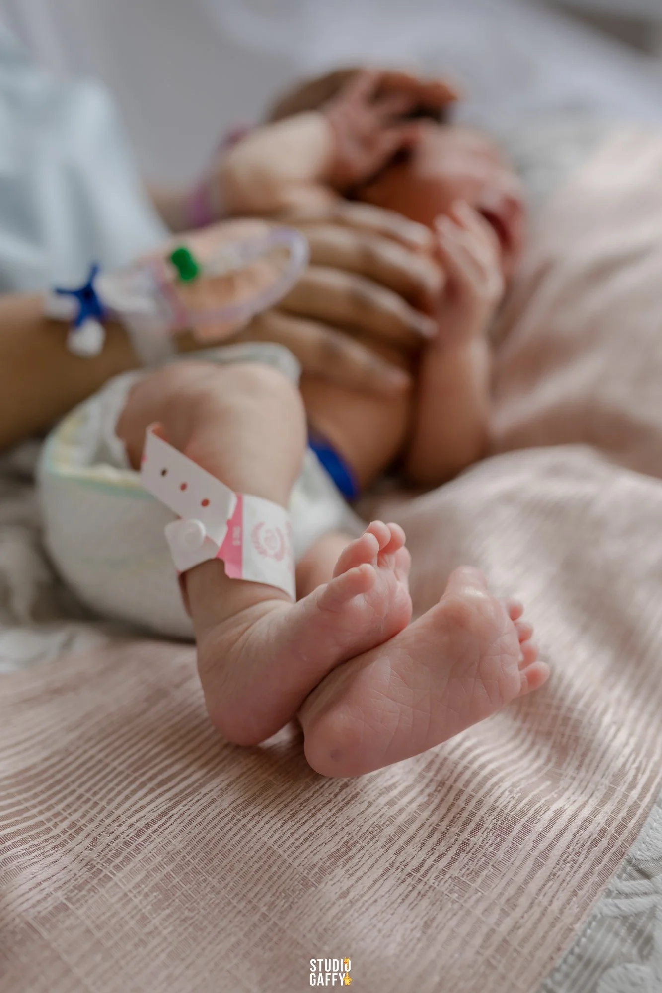 A newborn baby lying on a hospital bed with an IV line attached to the baby's leg, with an adult hand gently holding the baby's tiny hand.