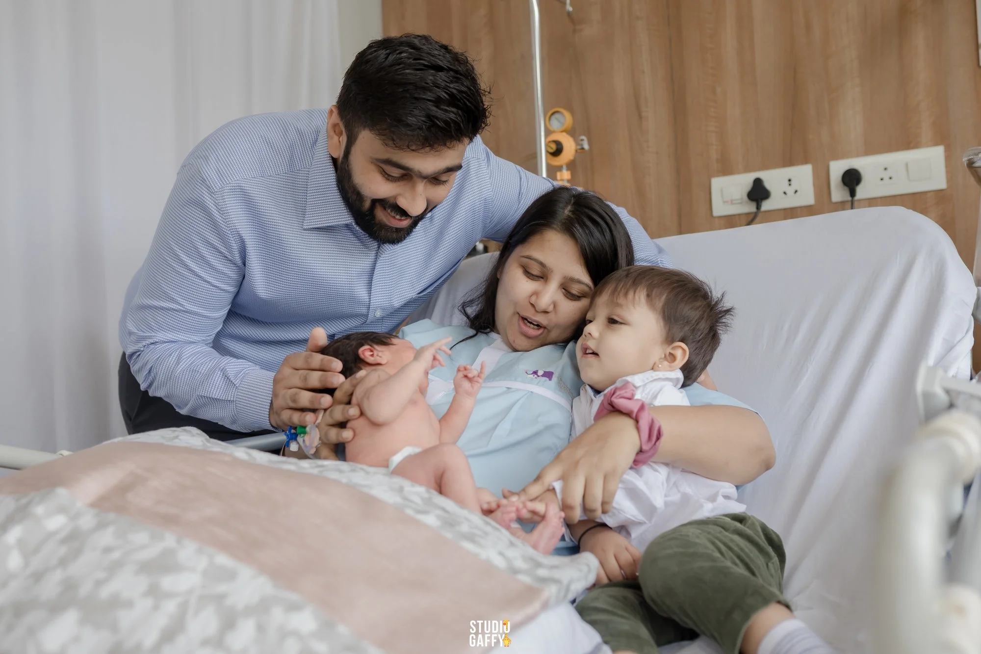 A family in a hospital room celebrating the birth of a baby. The mother is lying in a hospital bed holding the newborn, and the father and an older child are leaning over her, smiling and looking at the baby.