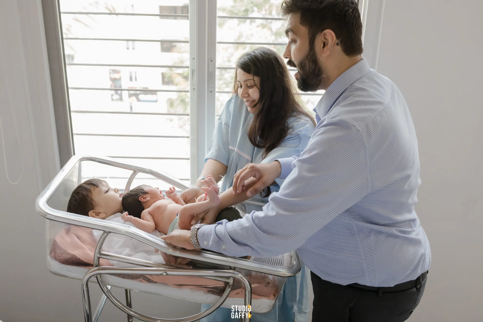 A man, woman, and two babies in a hospital nursery. The woman is feeding one baby while the man holds the other baby in a bassinet. Natural light from a window illuminates the scene.