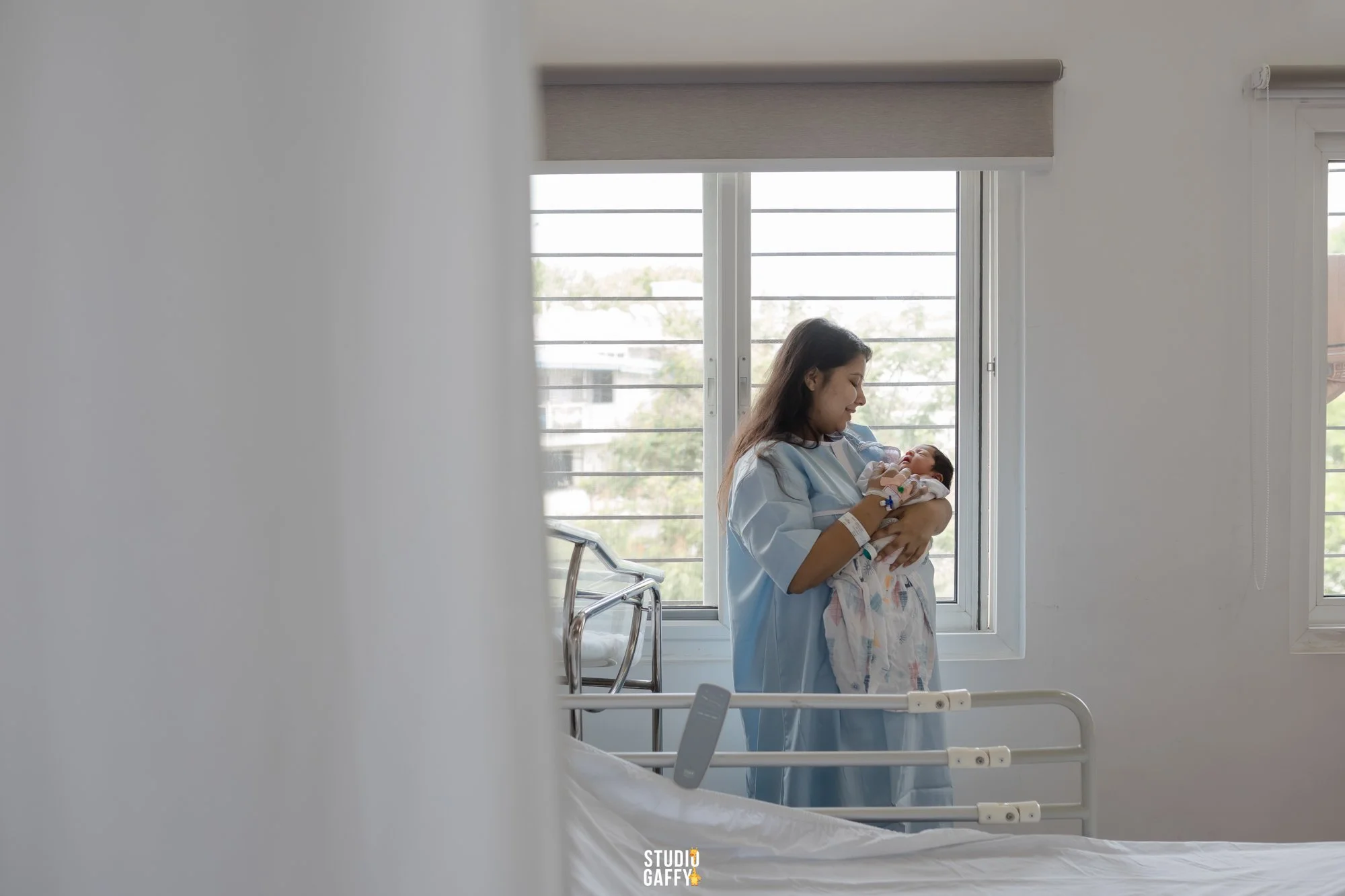 A woman in a hospital gown holding a newborn baby, standing by a window in a hospital room.