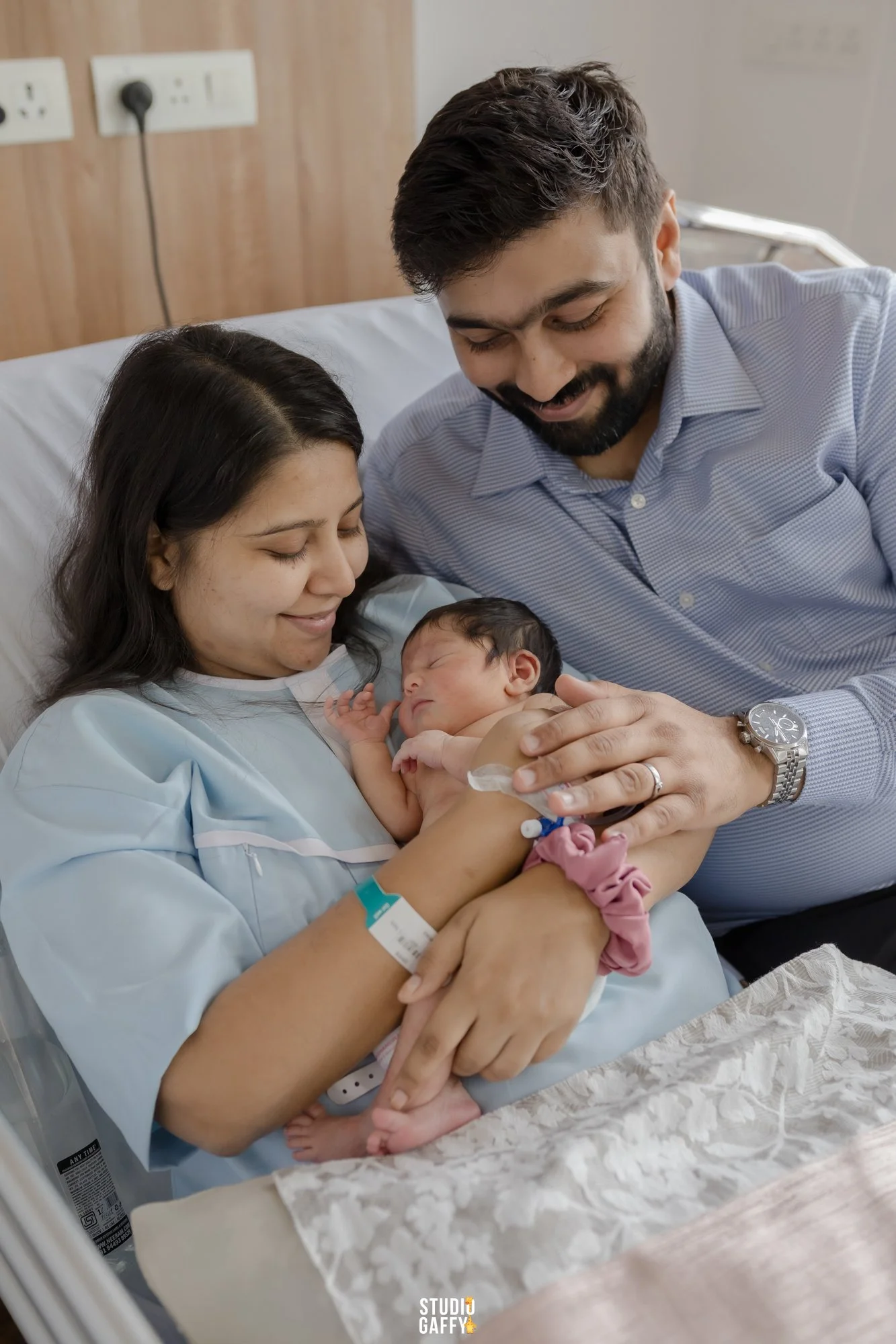 New mother in a hospital bed holding her newborn baby, with her partner leaning over and smiling.