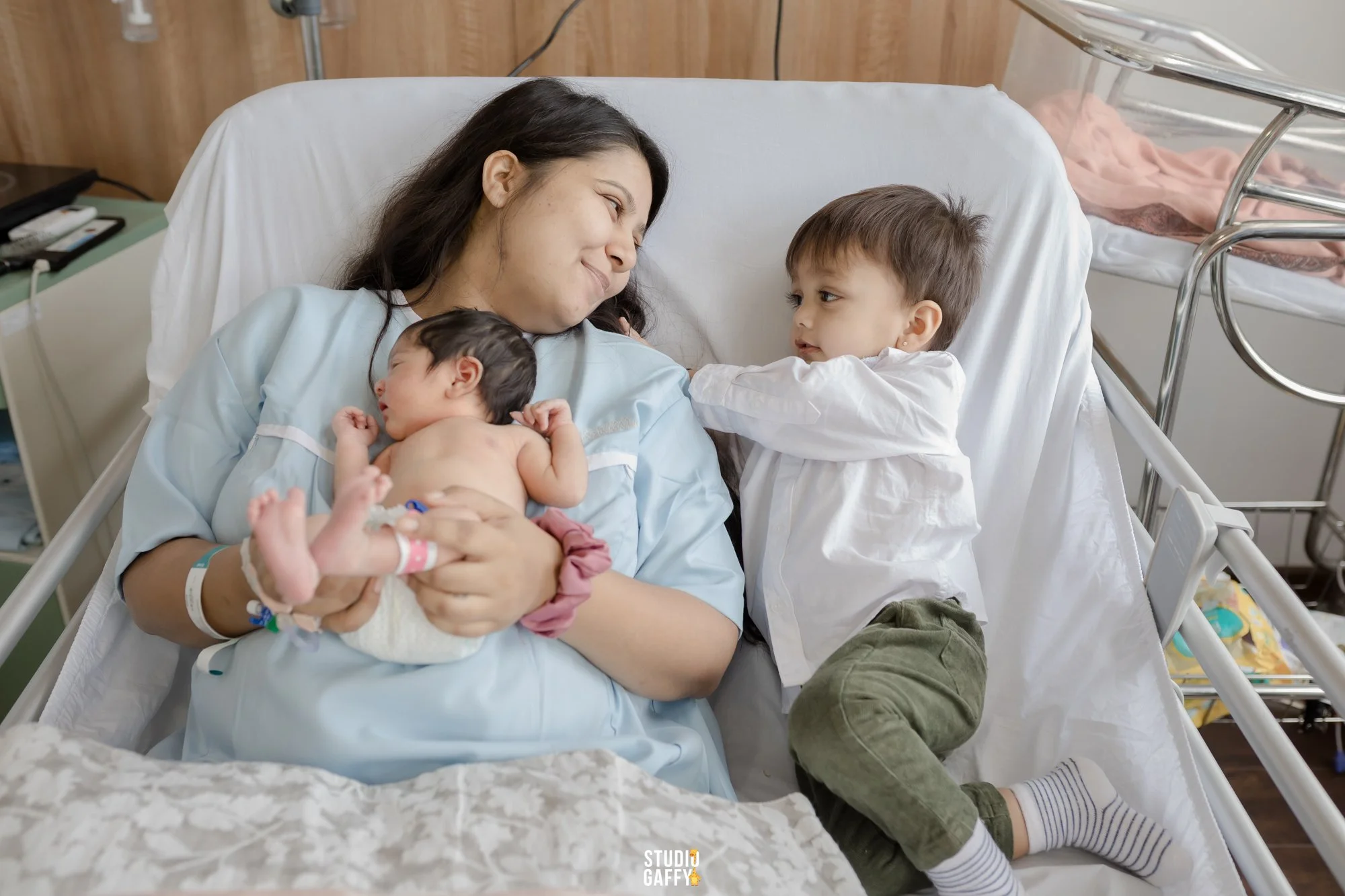 A woman lying in a hospital bed holding a newborn baby while a young boy laying next to her looks at them.