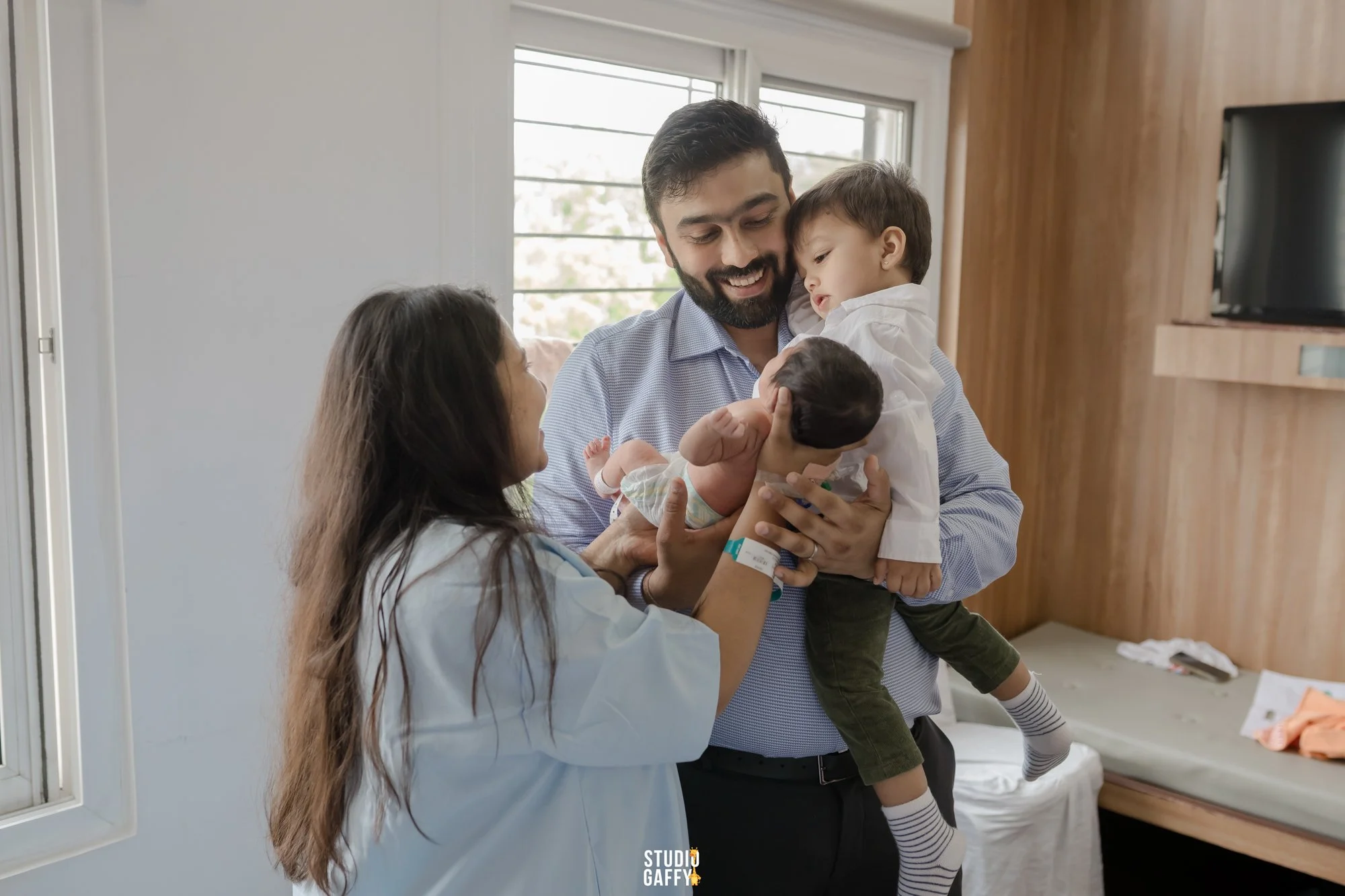A doctor holding a young boy, who is being examined by a nurse in a hospital room with large windows and wooden accents.