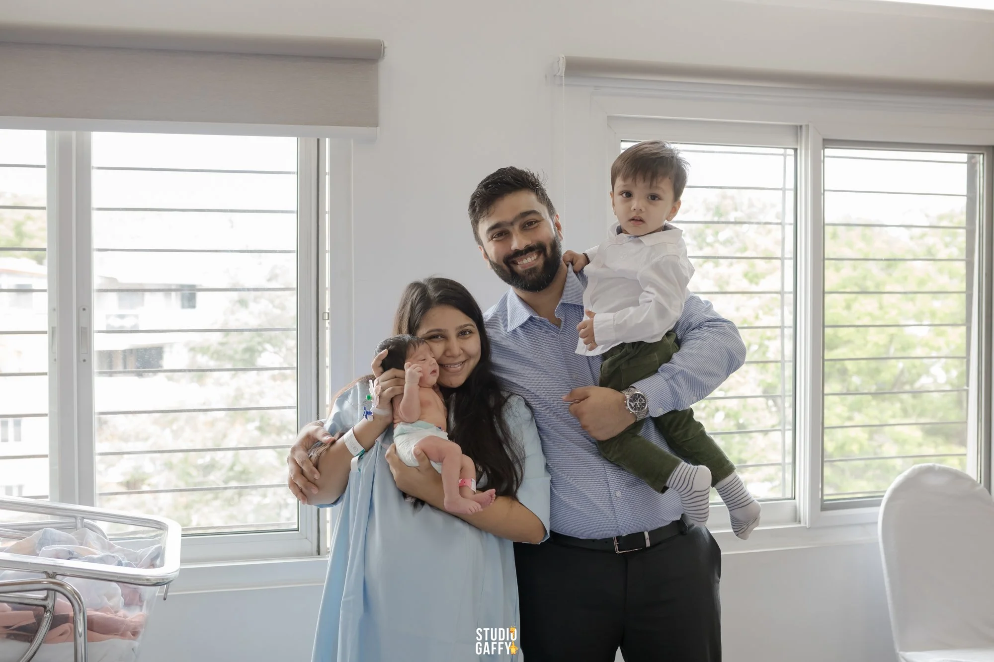 A family of four, including a mother, father, and two young children, standing together in a hospital room with large windows and hospital equipment, celebrating the birth of their newborn baby.
