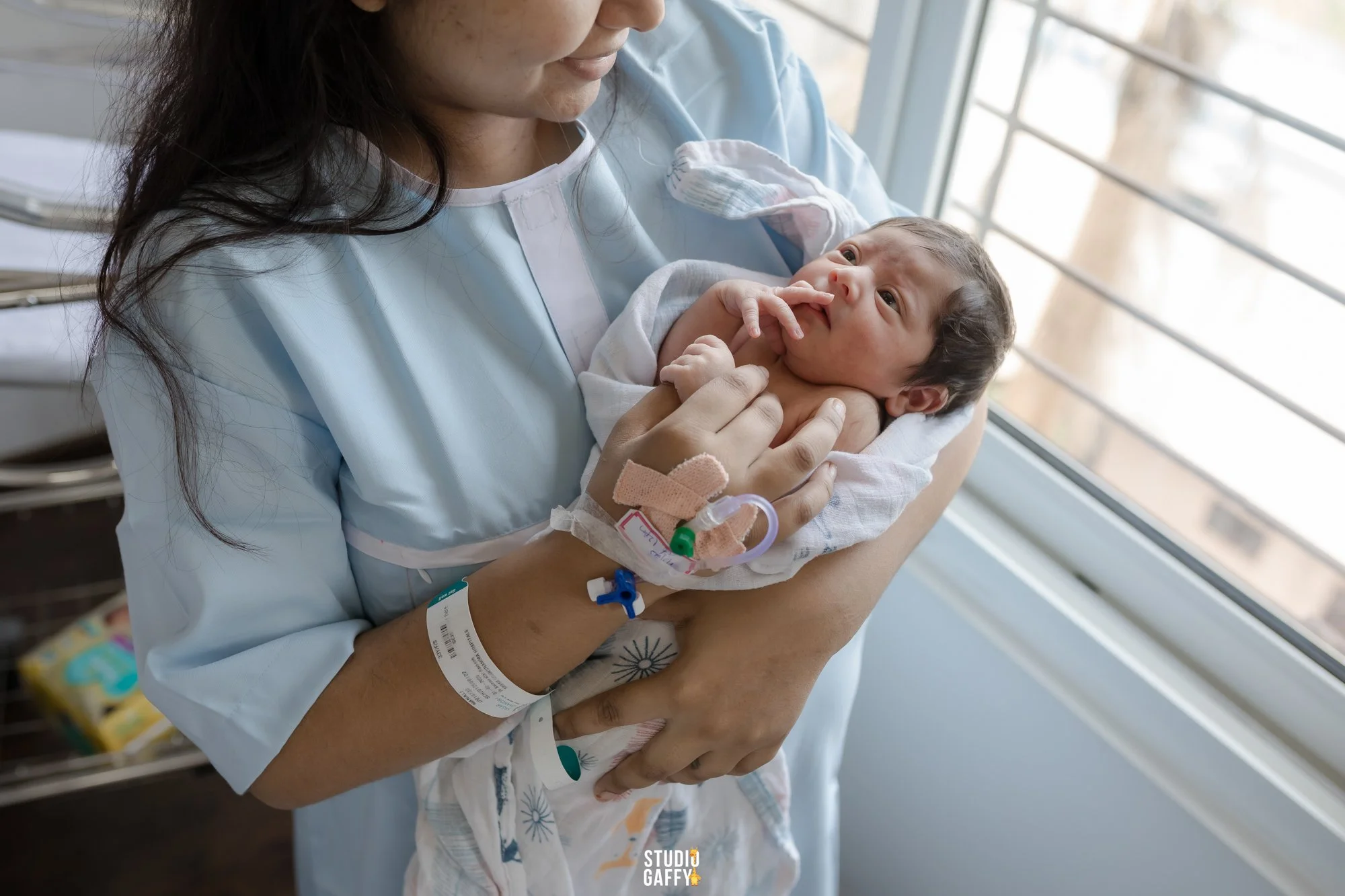 A woman in a hospital gown holding a newborn baby with medical tubes attached, sitting near a window in a hospital room.