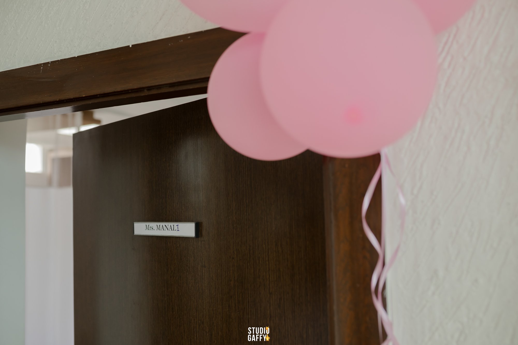 Pink balloons tied with pink ribbon, attached to a dark wooden structure with a white label reading 'Mrs. MANALI' in black text. The background appears to be an indoor setting with a window.