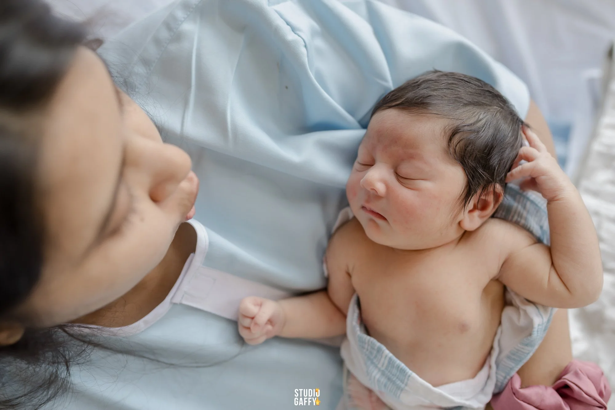 A woman lying down holding a newborn baby on her chest. The baby has dark hair and is resting with eyes closed, wearing a sleeveless outfit. The woman has long dark hair and is smiling softly, looking at the baby.