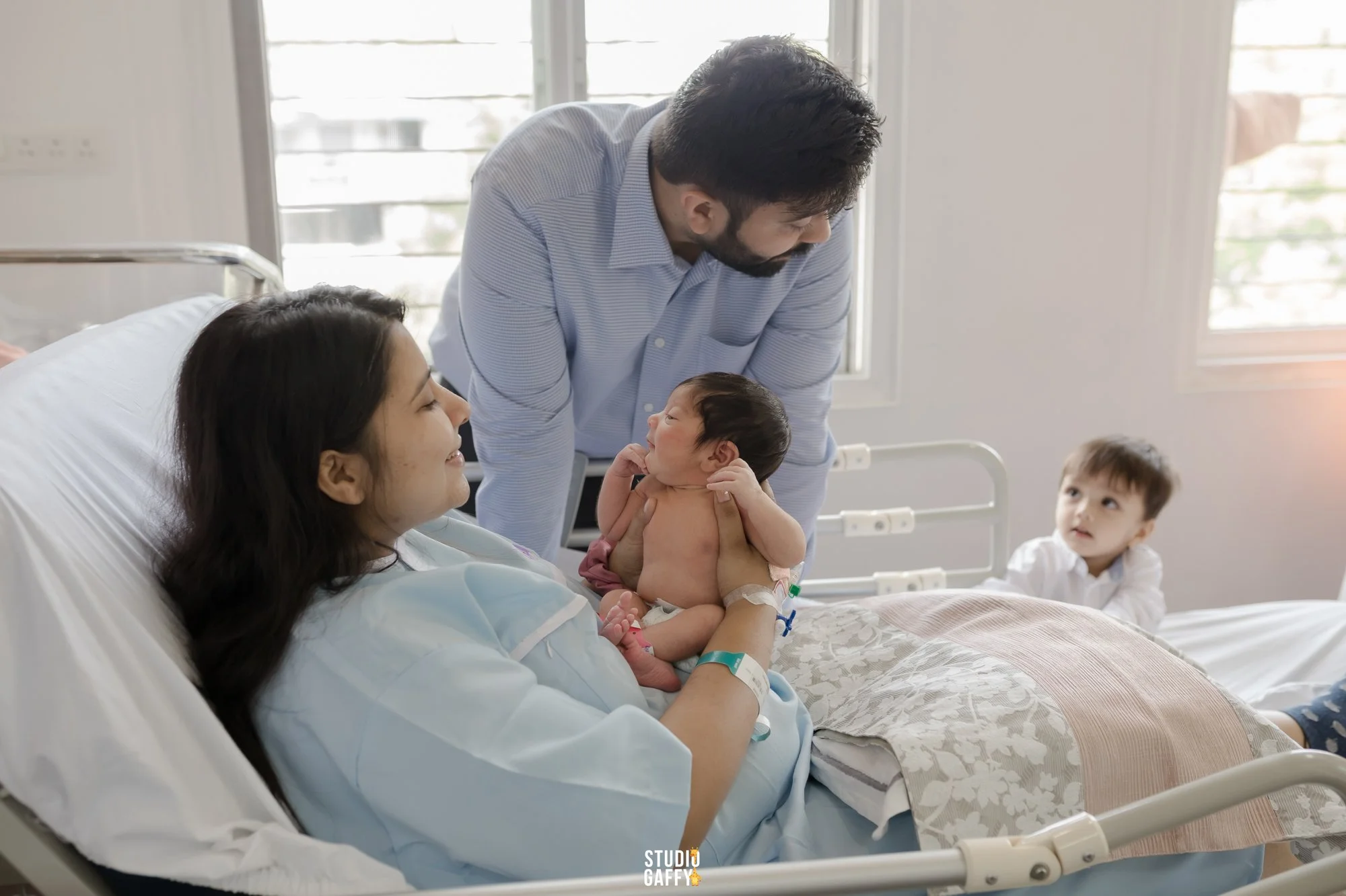 A woman in a hospital gown holding a newborn baby, a man in a striped shirt leaning over and smiling at the baby, and a young boy sitting on the hospital bed near the woman, all inside a hospital room with large windows.