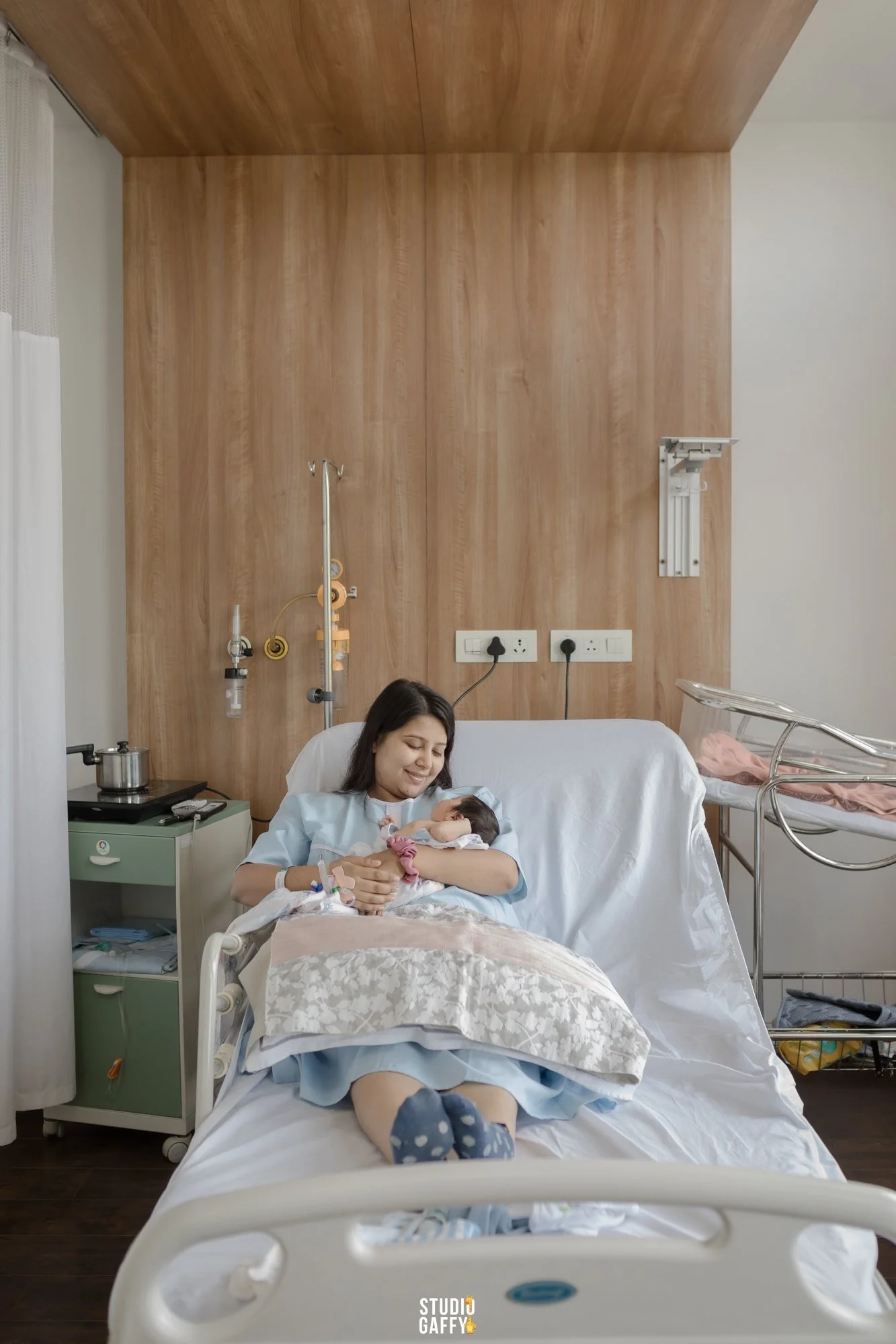 A woman in a hospital bed holding a newborn baby, smiling and looking at the baby. The room has wooden wall panels, medical equipment, and a hospital tray table.