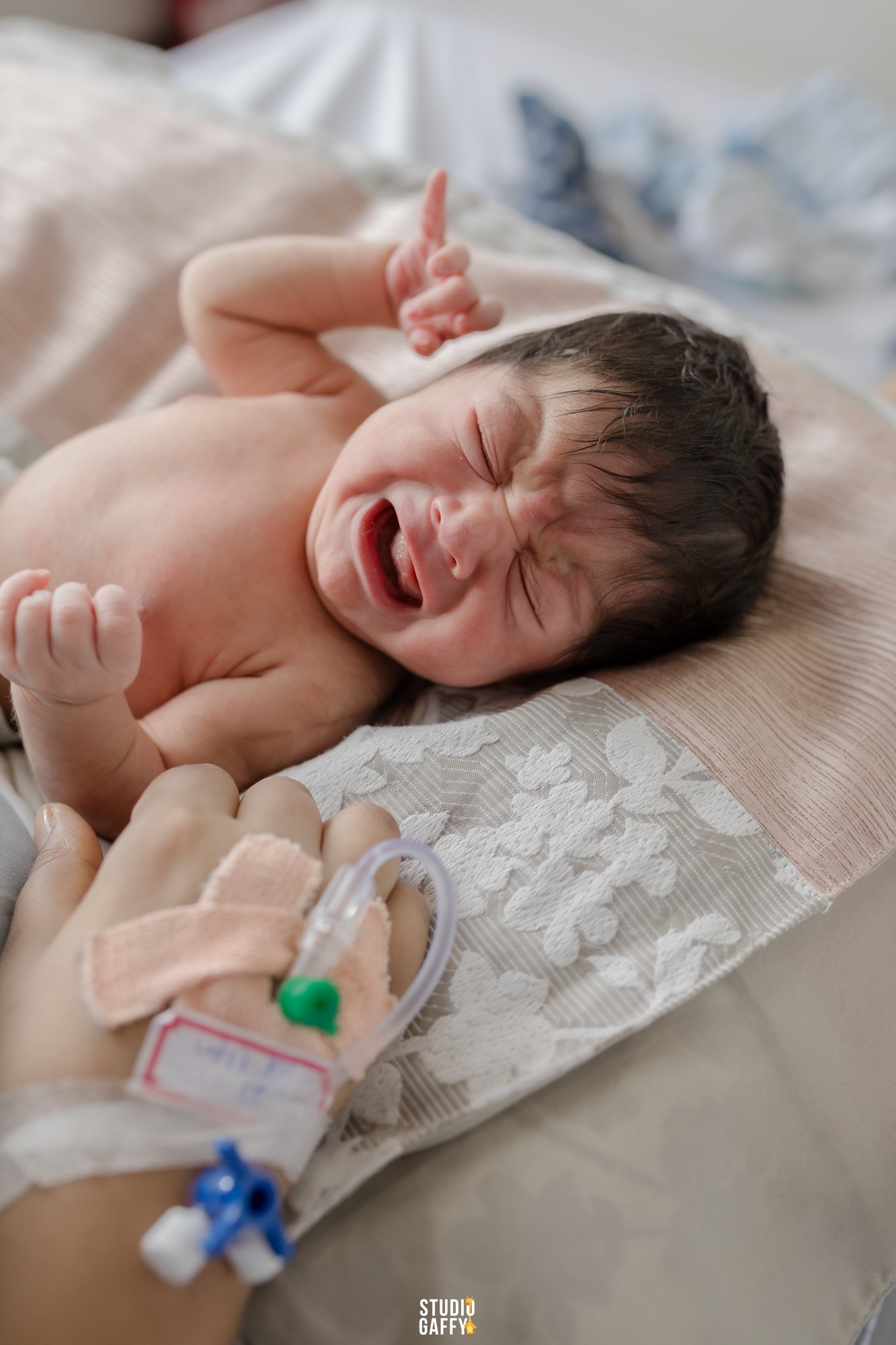 A newborn baby lying on a bed, smiling and holding up one finger, with medical monitoring equipment attached to their hand.