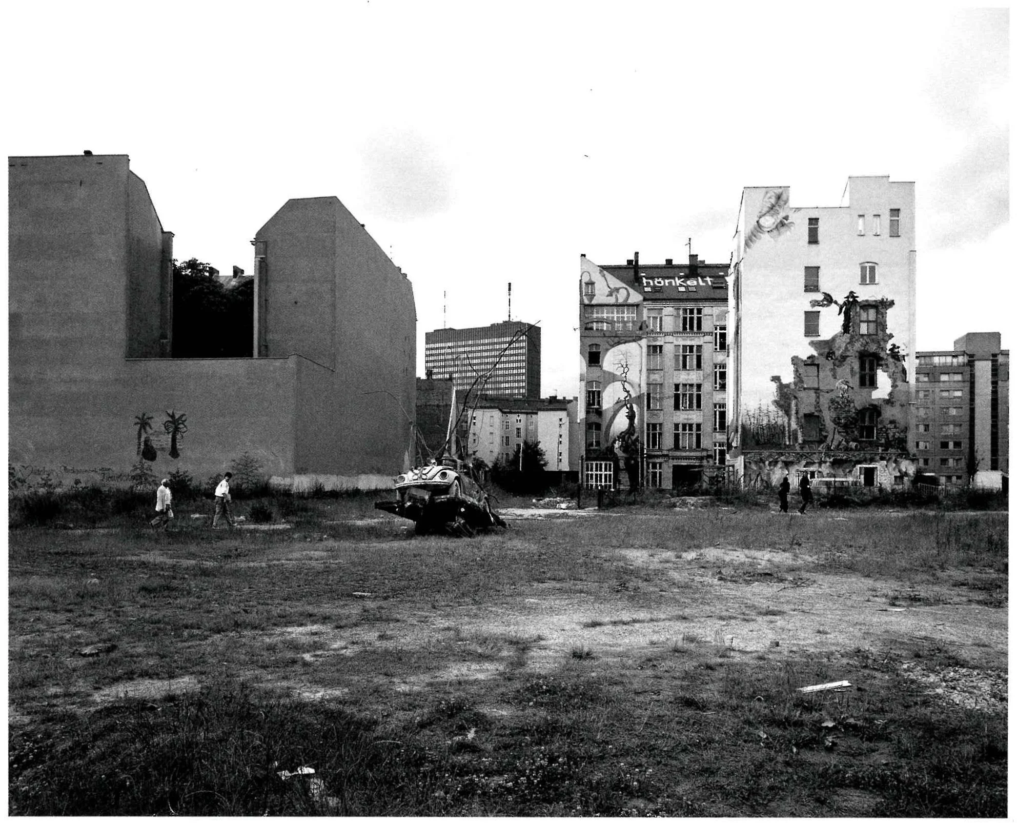 A black-and-white photo of an urban vacant lot with dilapidated buildings, graffiti, and a wrecked car. Pedestrians are walking through the area with tall buildings in the background.