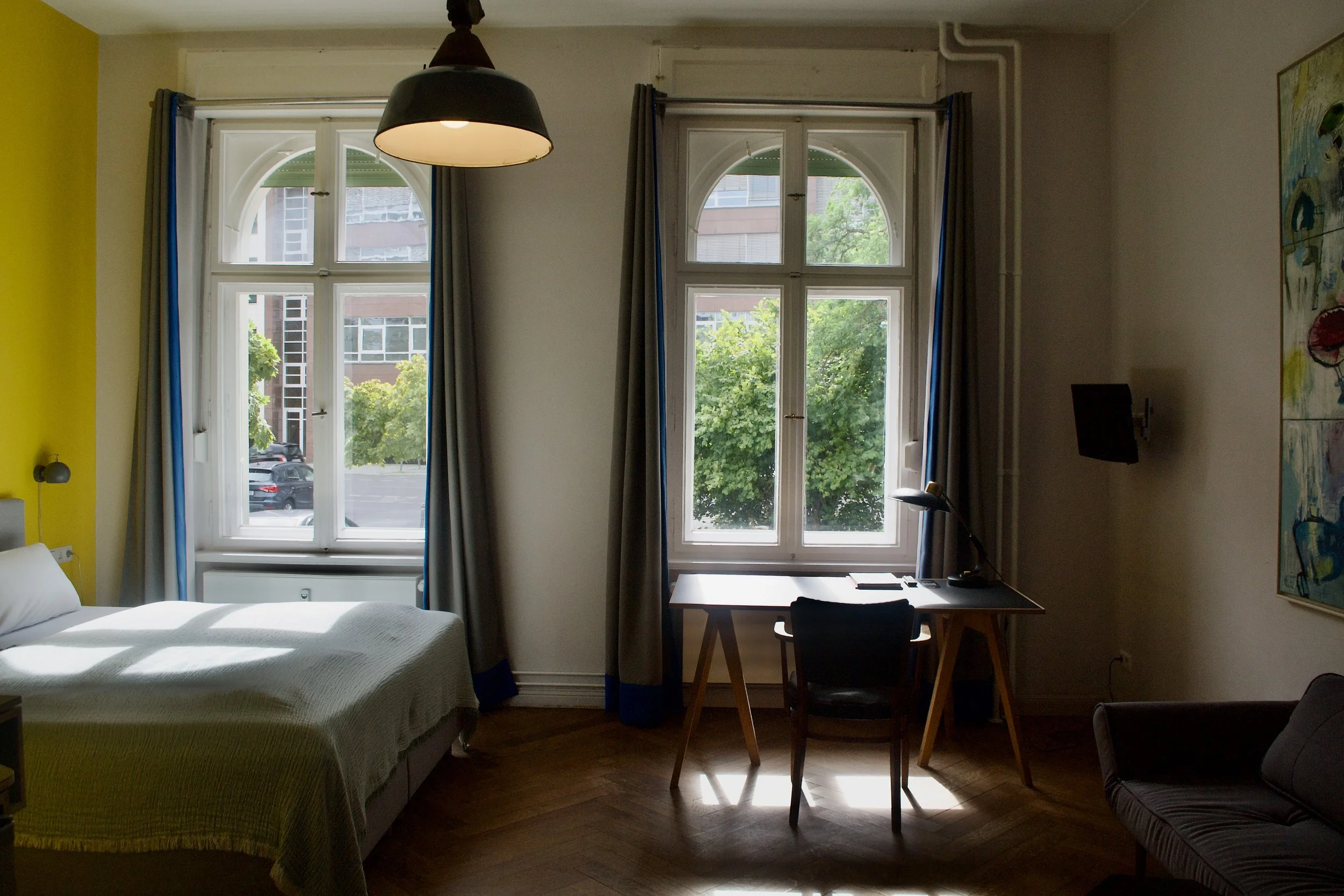 A cozy bedroom with a bed, large windows, a desk with a chair, and a piece of colorful artwork on the wall.