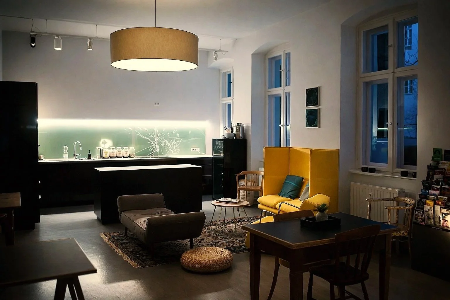 Cozy living room with large windows, yellow high-back armchair, wooden coffee table, sofas, and a kitchen area with a backsplash, pendant light, and various kitchen appliances.