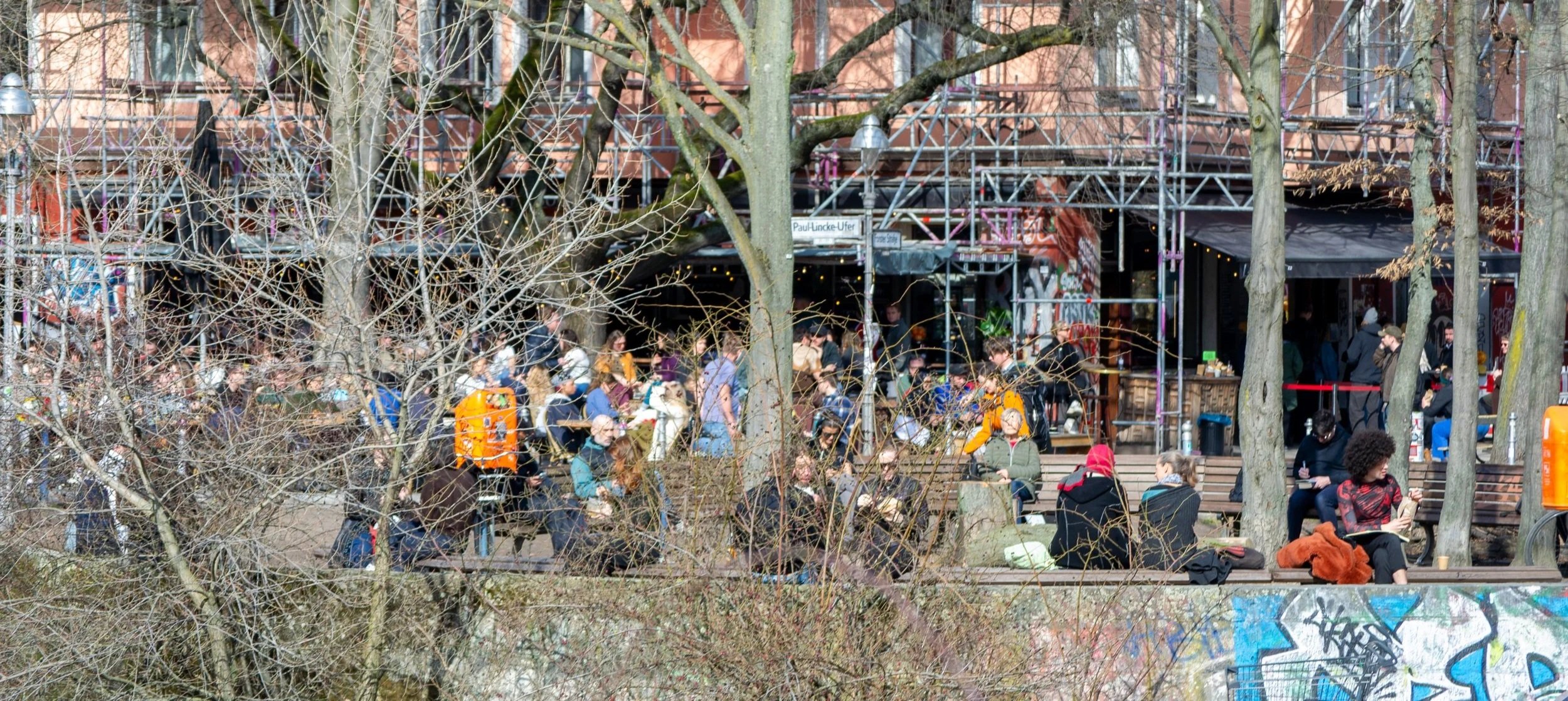 People gathered outdoors in an urban space with trees, benches, and a graffitied wall, some sitting, some standing, and a building with scaffolding in the background.
