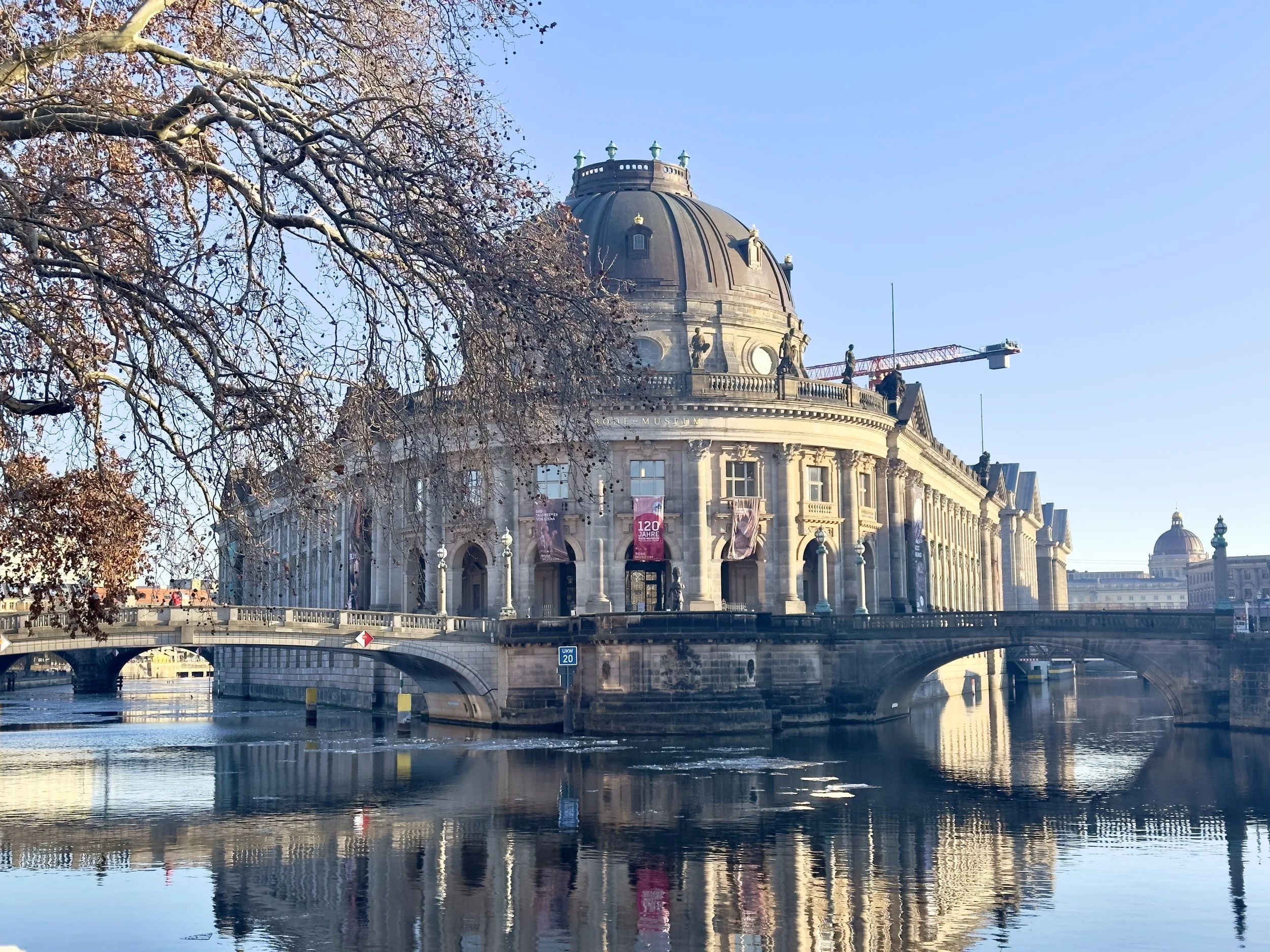 Historical building with a dome, reflected in the river, with leafless tree branches on the left and a construction crane on the right, under a clear blue sky.