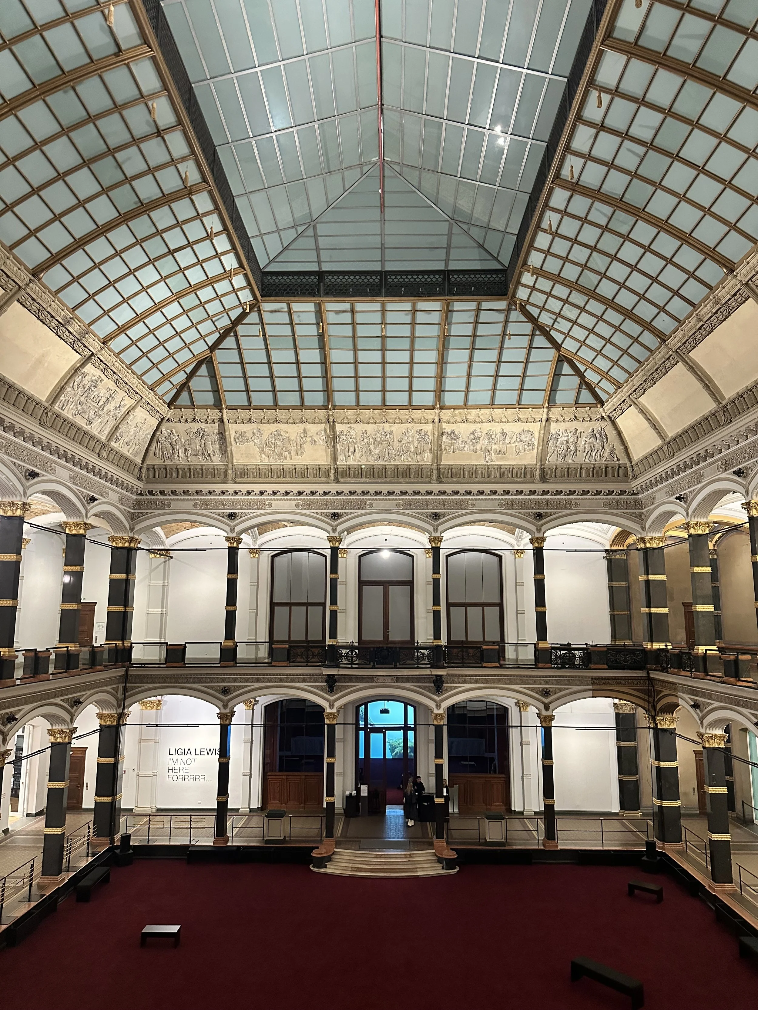 Interior view of a historic building with a large glass ceiling, multiple floors with arched doorways, decorative columns with gold accents, and a maroon carpeted floor.