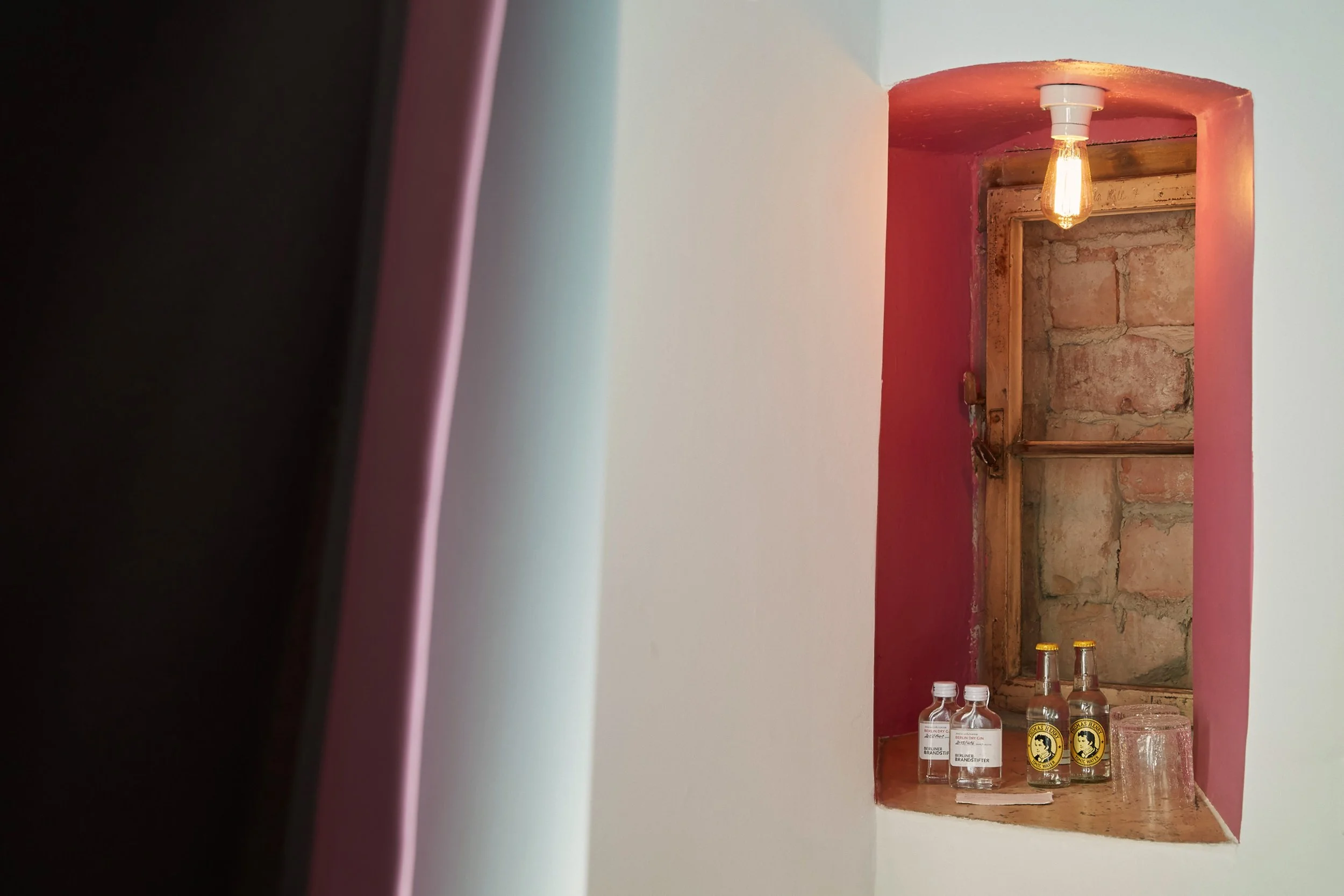 Narrow wall niche with exposed brick and pink-painted wall, containing bottled beverages, a glass, and a lightbulb.