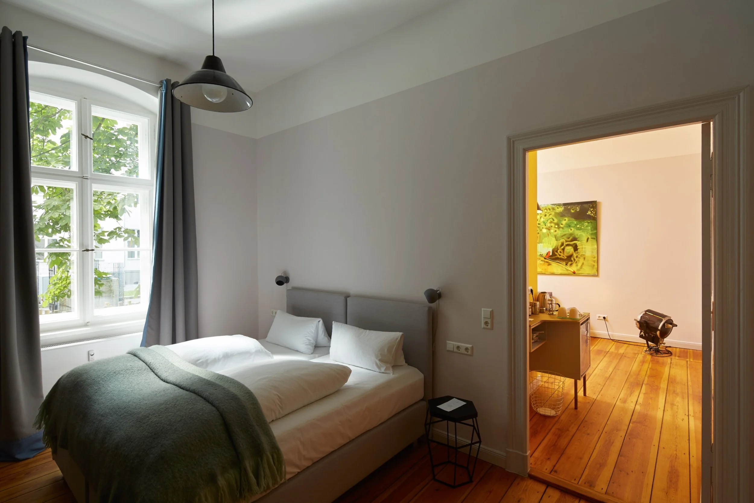 Bedroom with white walls, large window with blue curtains, bed with white bedding and green throw blanket, small black side table, and open door leading to a room with wooden floor, artwork, coffee station, and a lamp