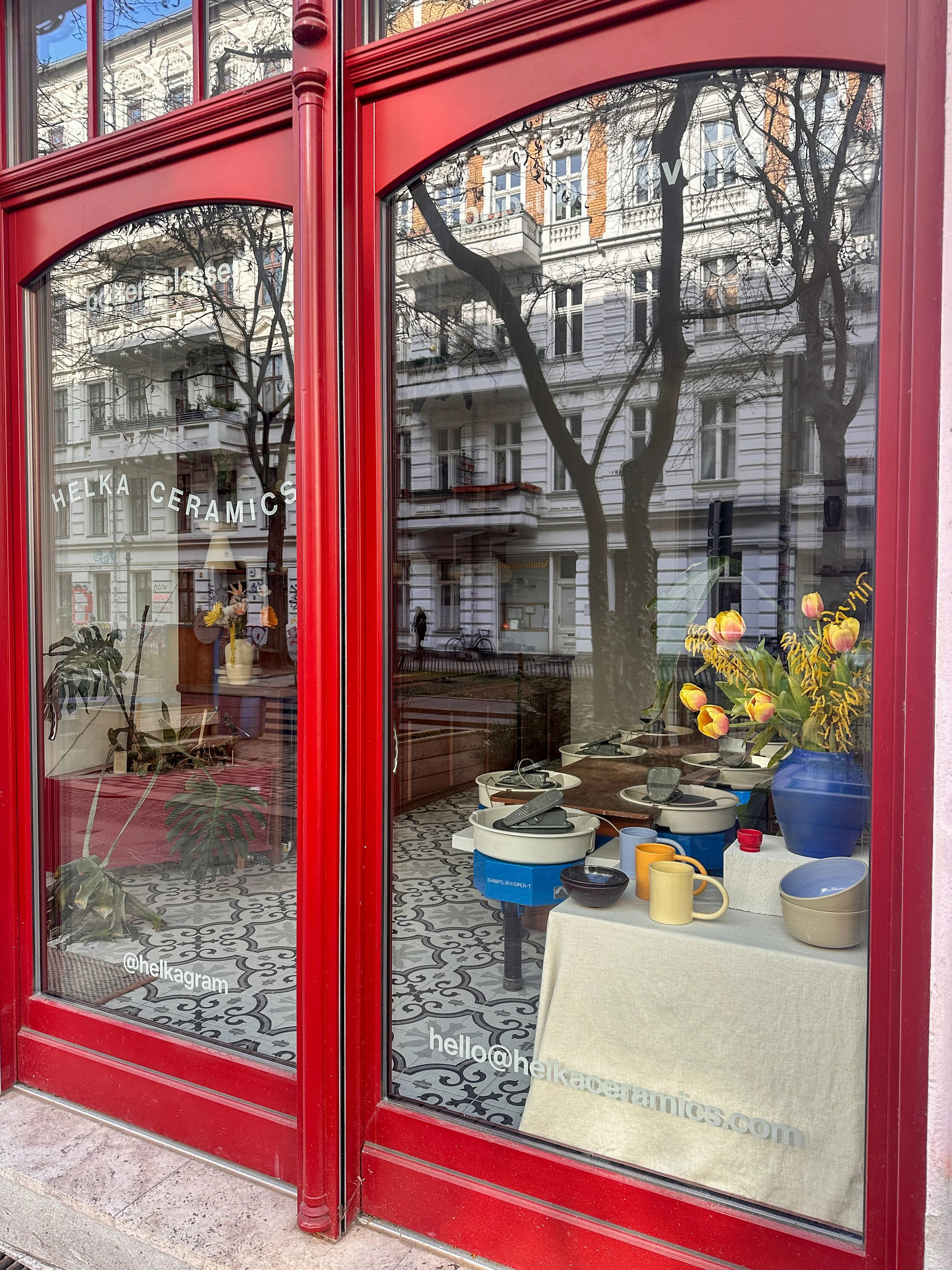 View through a large red-framed window showing a ceramic shop interior with pottery, cups, bowls, flowers, plants, and decorative items on display inside. The shop window reflects buildings and trees outside.